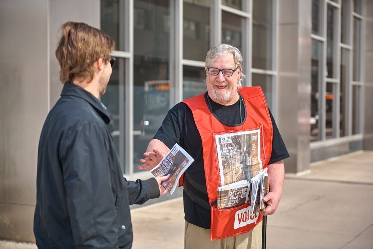 A man wearing a red volunteer vest handing a pamphlet to a young man outside a building.
