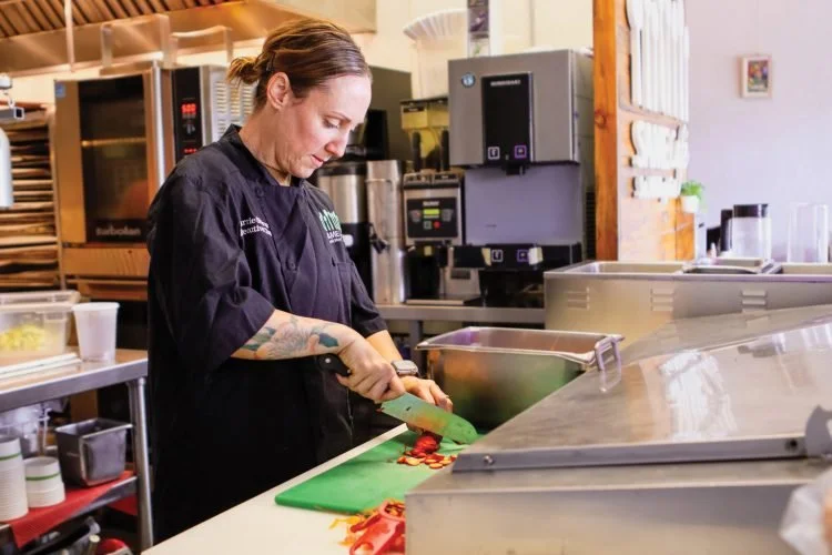 A woman in a black chef's coat is chopping red bell peppers on a green cutting board in a commercial kitchen.