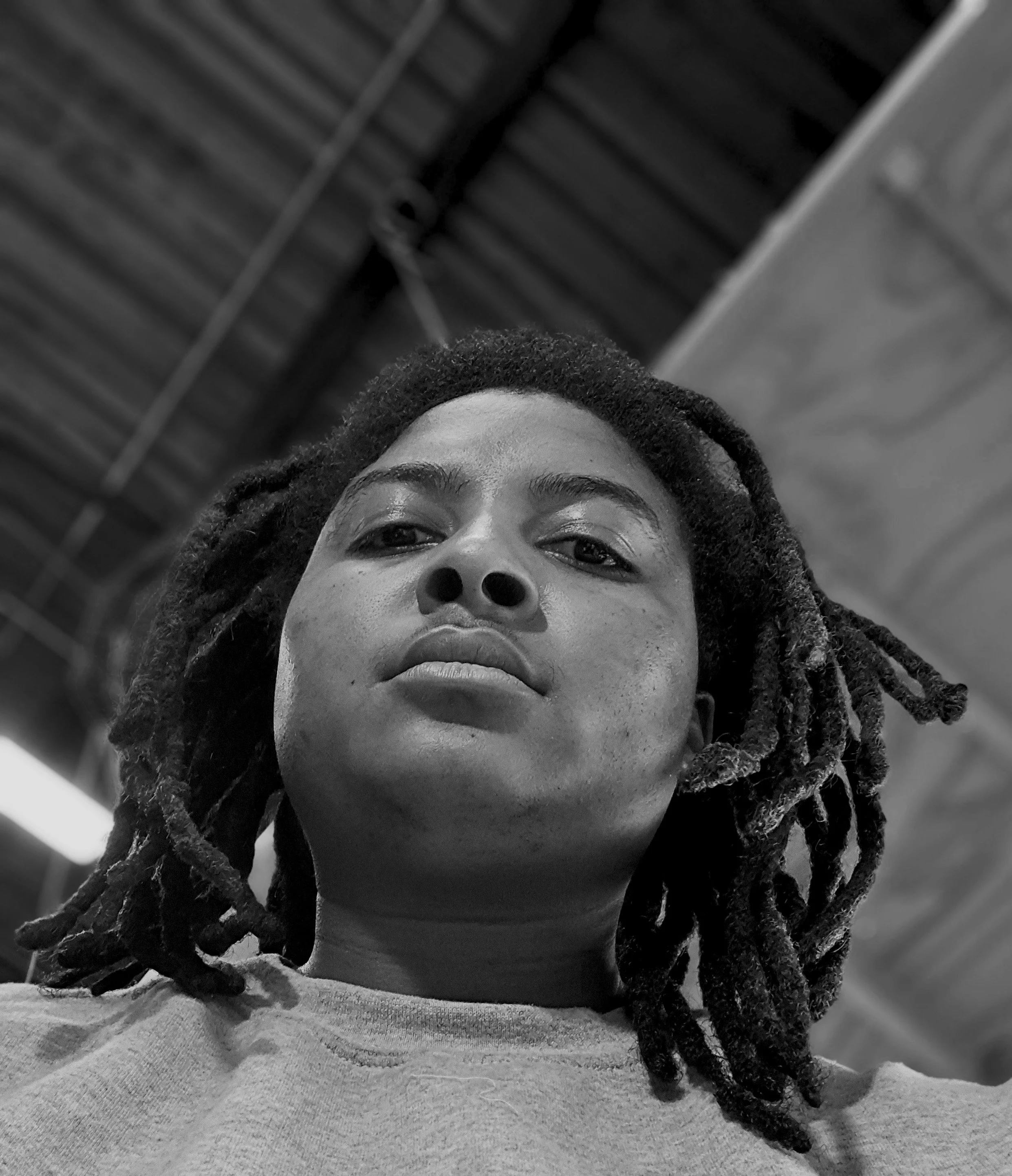 Close-up black and white photo of the artist, K.Gilbert, with dreadlocks, looking down at the camera with a confident expression, an industrial ceiling in the background.