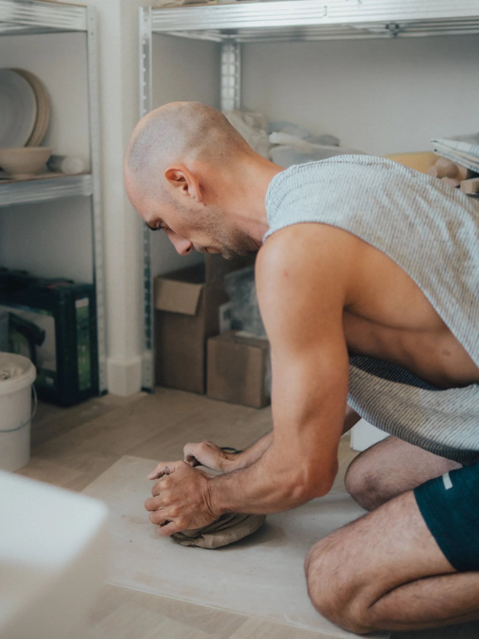 A man with a shaved head and wearing a sleeveless shirt kneels on the floor, leaning forward with his hands on a cloth or bag. He appears to be focused on something in front of him in a room with metal shelving and various storage containers.