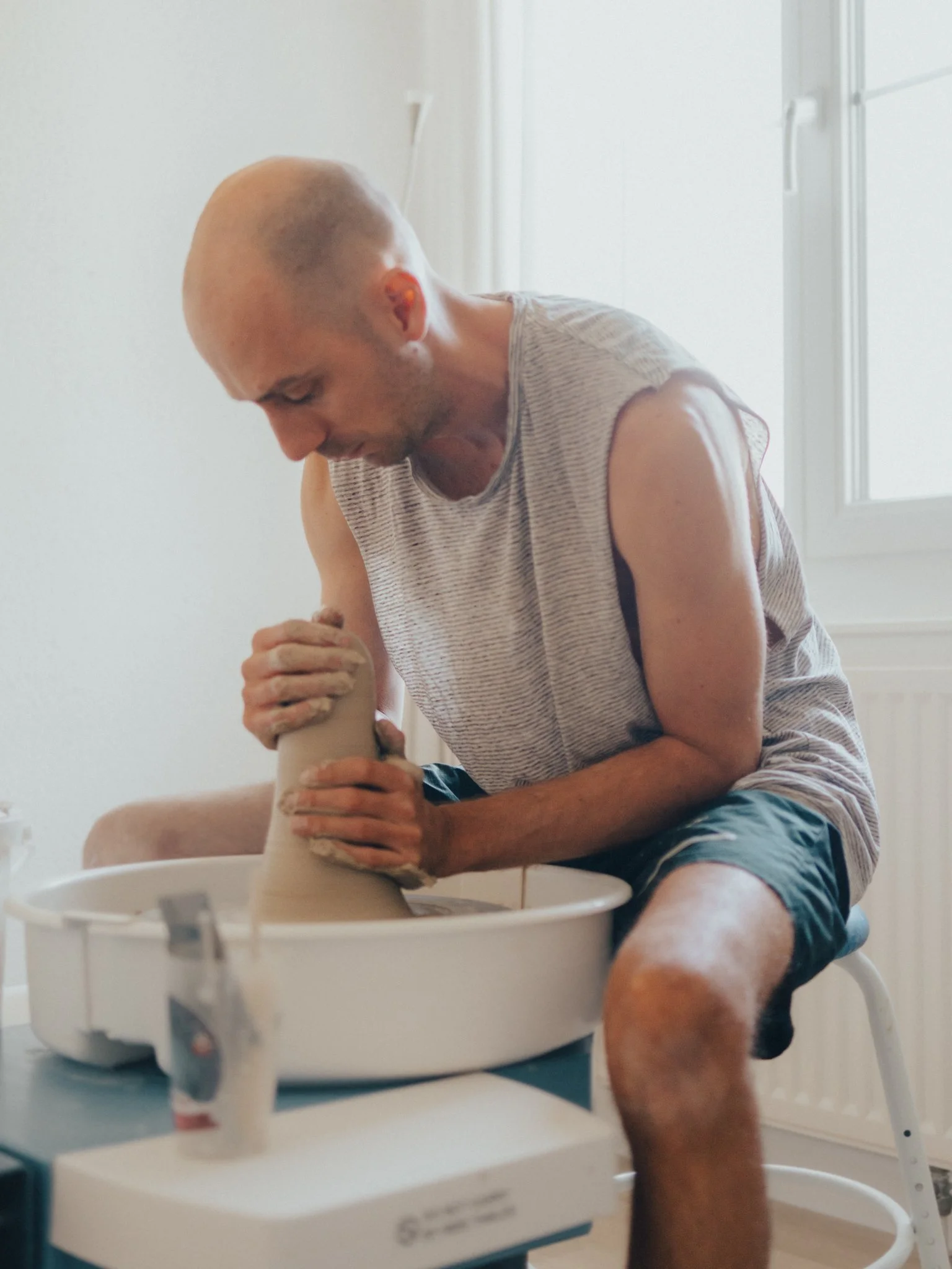 A man with a bald head and a beard, wearing a striped sleeveless shirt and shorts, is sitting on a white chair making a pottery piece on a pottery wheel in a bright room.