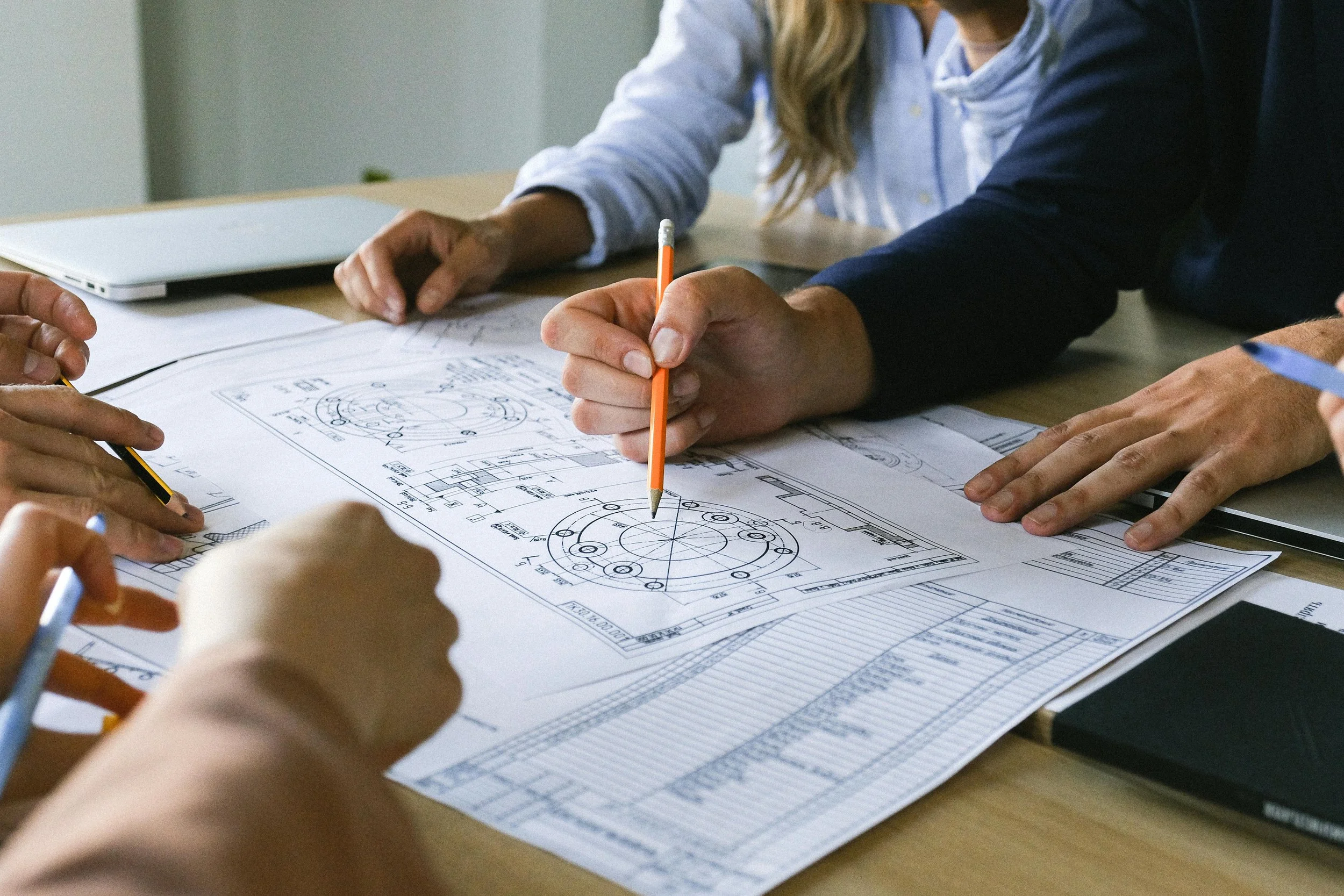 Group of people working on technical blueprints and engineering plans at a desk, with a laptop nearby.