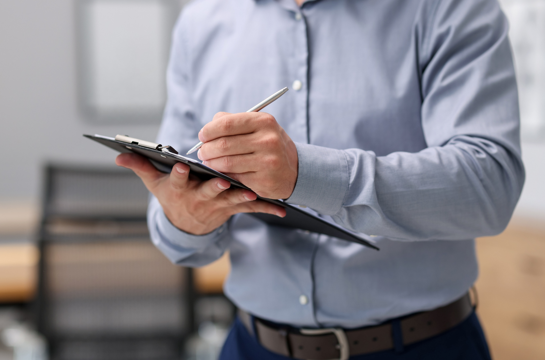 Person wearing a blue button-up shirt writing or taking notes on a clipboard with a pen, in an office setting.