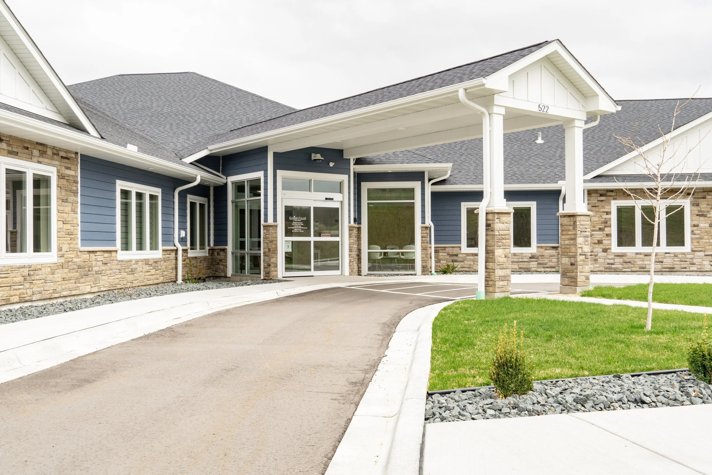 A modern building with a blue exterior, stone accents, and a covered entrance supported by four stone columns. There is a curved driveway and small landscaped areas with grass, small bushes, and a young tree.