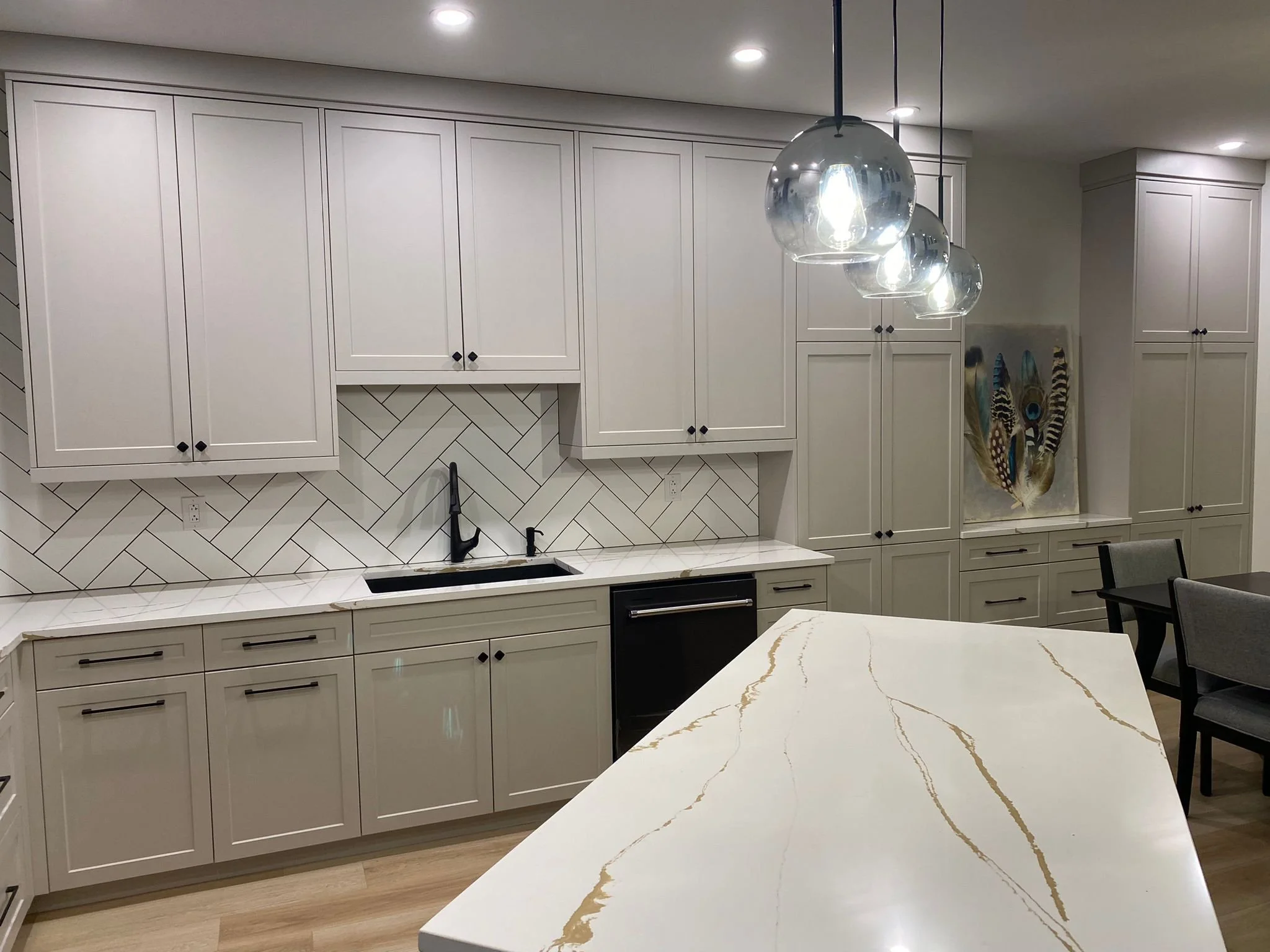 Modern kitchen with white cabinets, black hardware, a black sink and faucet, and a white countertop with gold veining. Pendant lights hang above a kitchen island, and there is artwork with feathers on the wall.