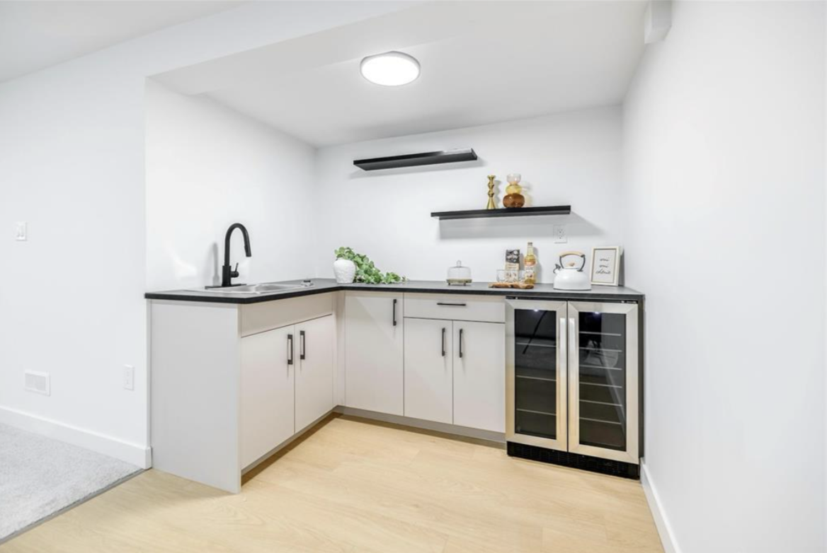 Small modern kitchenette with white cabinets, black countertop, black faucet, two shelves with decorative items, and a mini refrigerator with a glass door.
