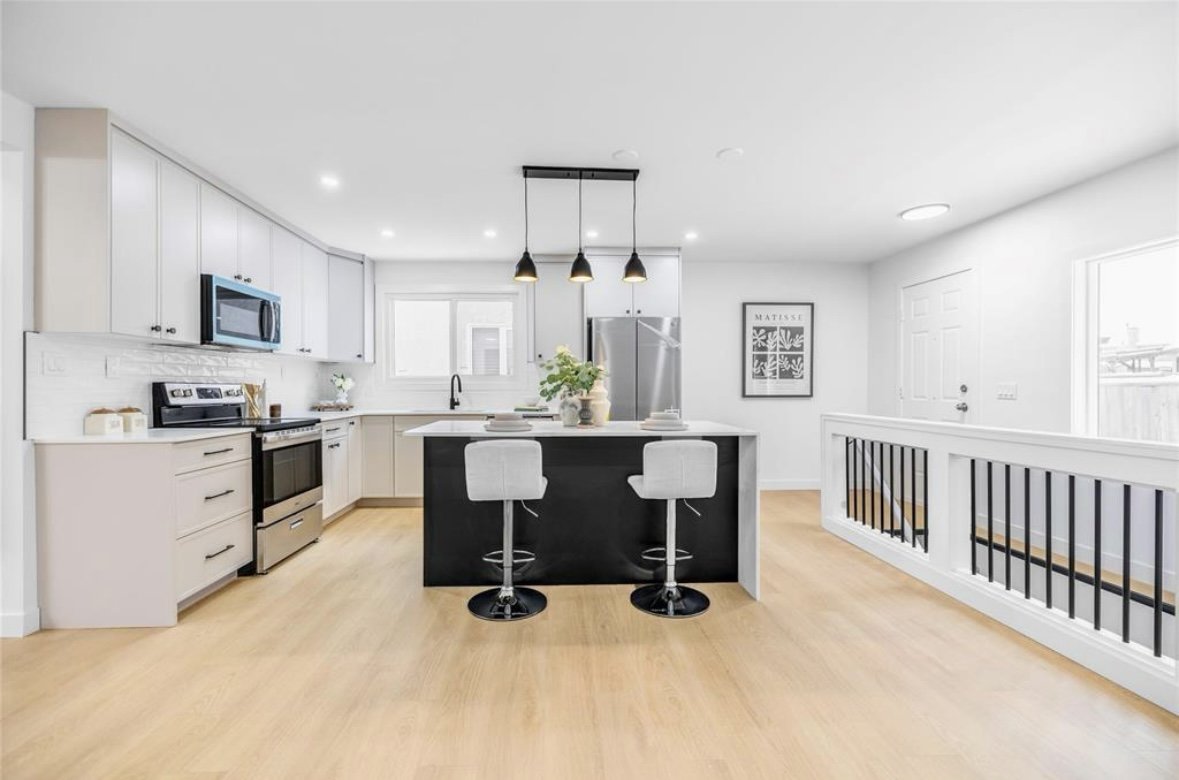 Modern kitchen with white cabinets, a black island with two bar stools, stainless steel appliances, and light wood flooring.