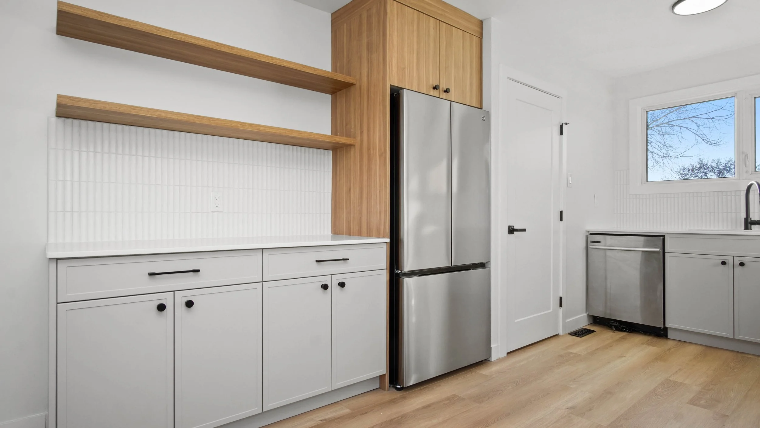 Modern kitchen with white cabinets, wooden open shelves, a stainless steel refrigerator, and a window showing trees outside.