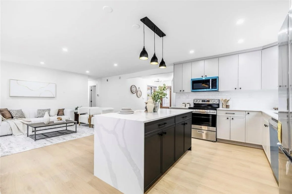 Open-concept kitchen and living room with white and black cabinets, stainless steel appliances, a kitchen island with a plant, and a white seating area with a coffee table