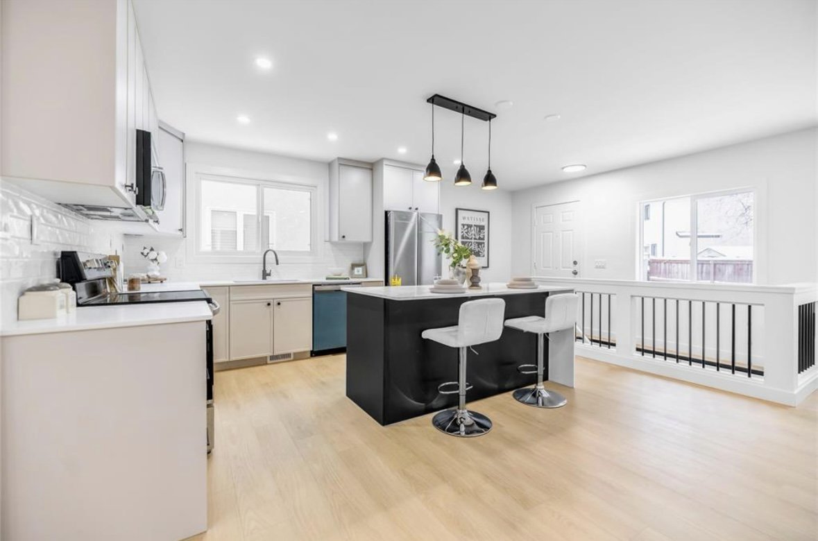Modern kitchen with white cabinetry, a black island with white stools, stainless steel appliances, and light hardwood floors.