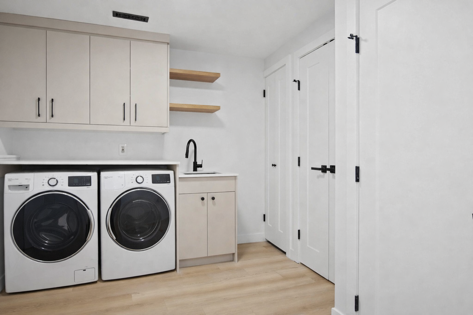 A laundry room with a white washer and dryer set, white cabinets, open wooden shelves, a small sink with a black faucet, and white louvered doors.