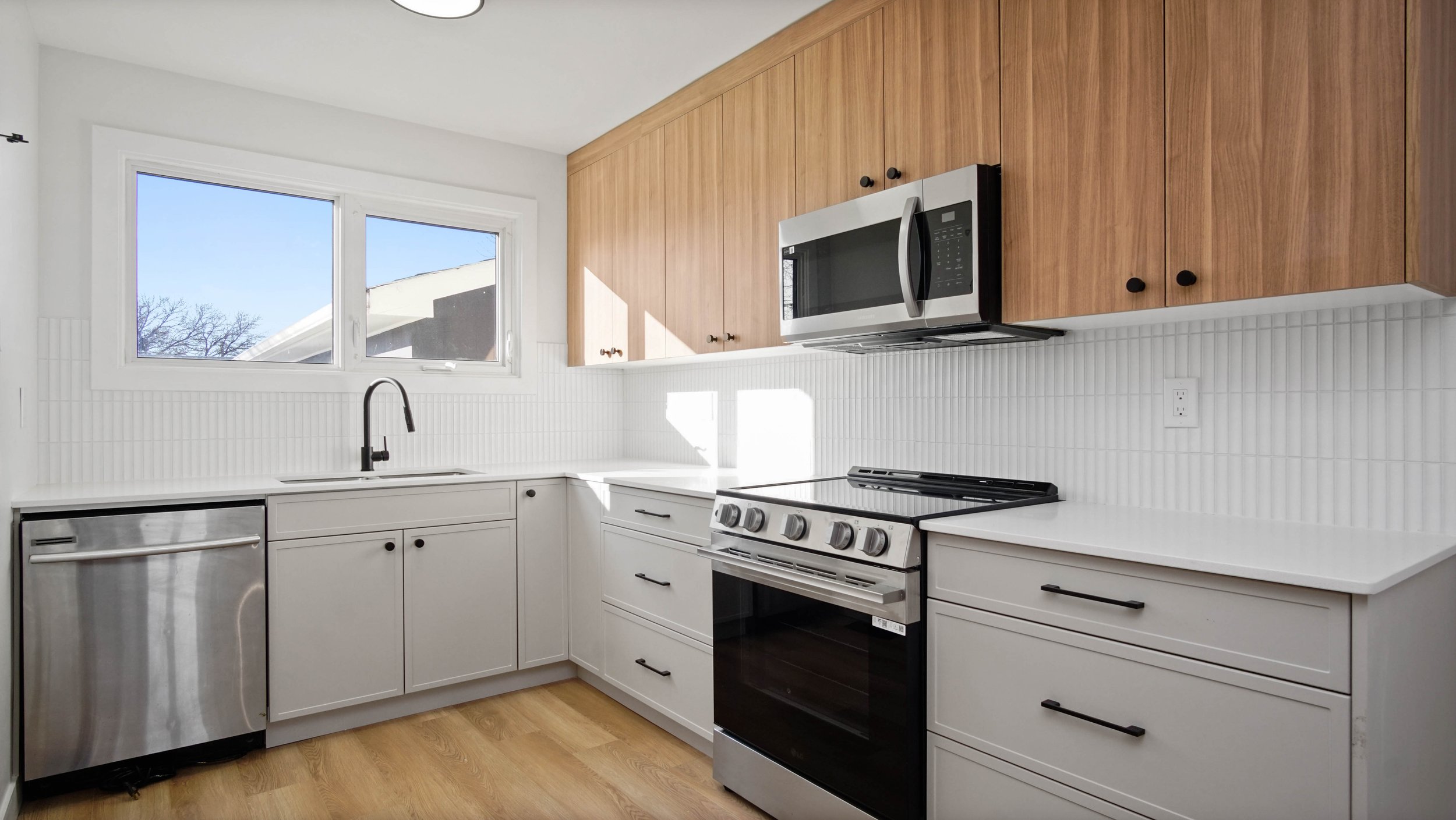 Modern kitchen with white cabinets, a stainless steel stove, microwave, and small refrigerator, wooden upper cabinets, white beadboard backsplash, window above sink, and natural light.