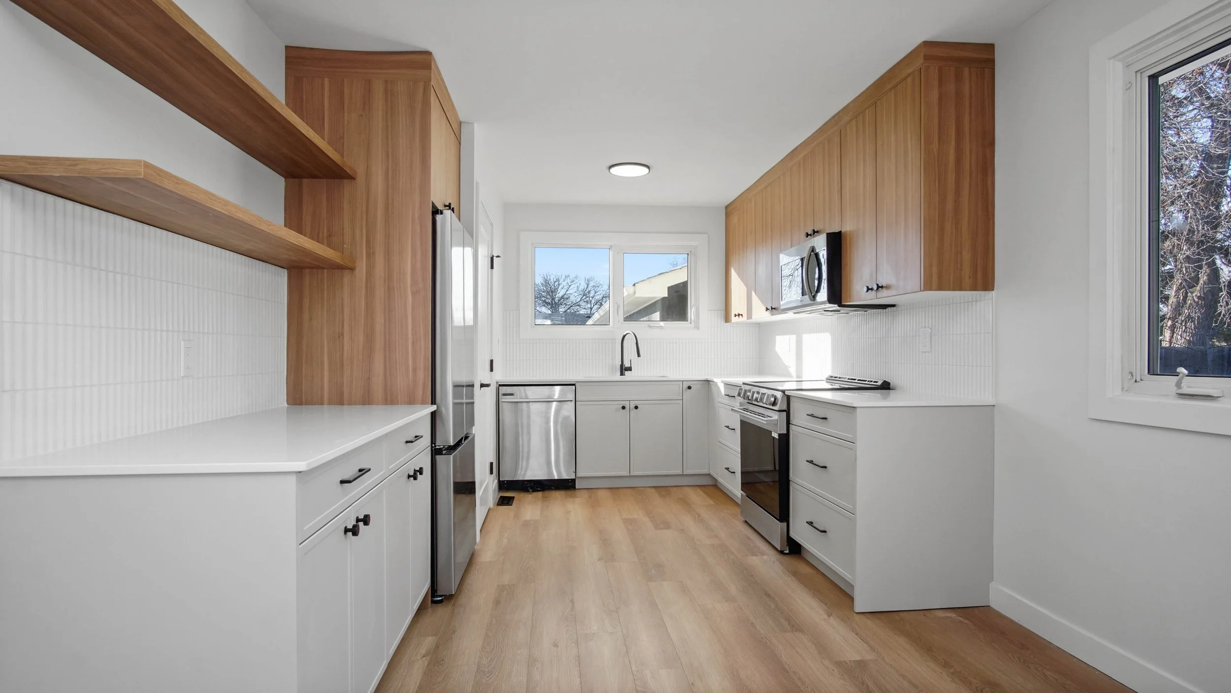 Modern kitchen with wooden upper cabinets, white lower cabinets, kitchen appliances, and hardwood flooring, with natural light coming through windows.