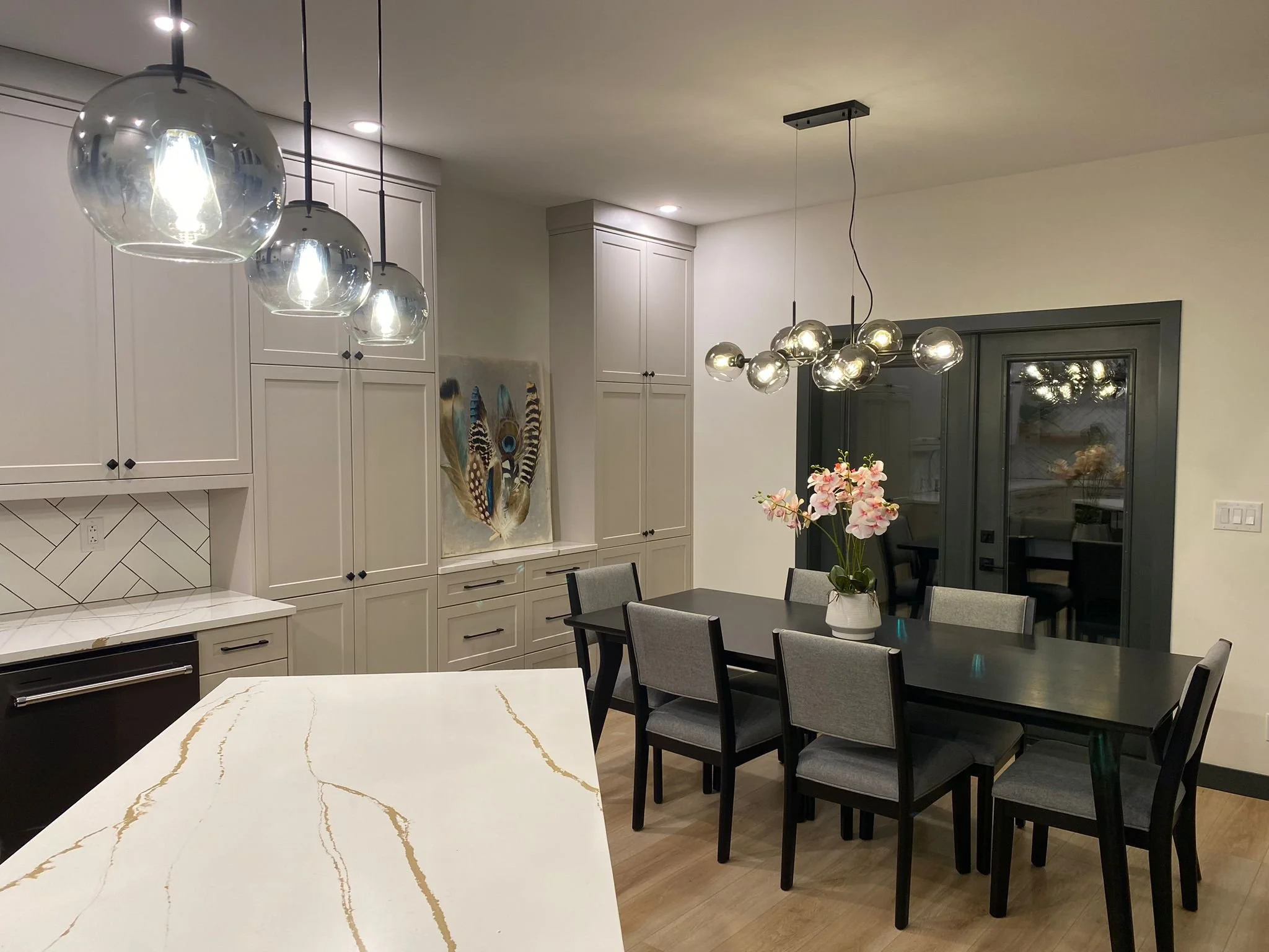 A modern dining room with a black table, gray upholstered chairs, and a vase of pink orchids. The room features white cabinetry on the left, a framed artwork of a peacock feather on the wall, and a contemporary chandelier with glass globes. There are