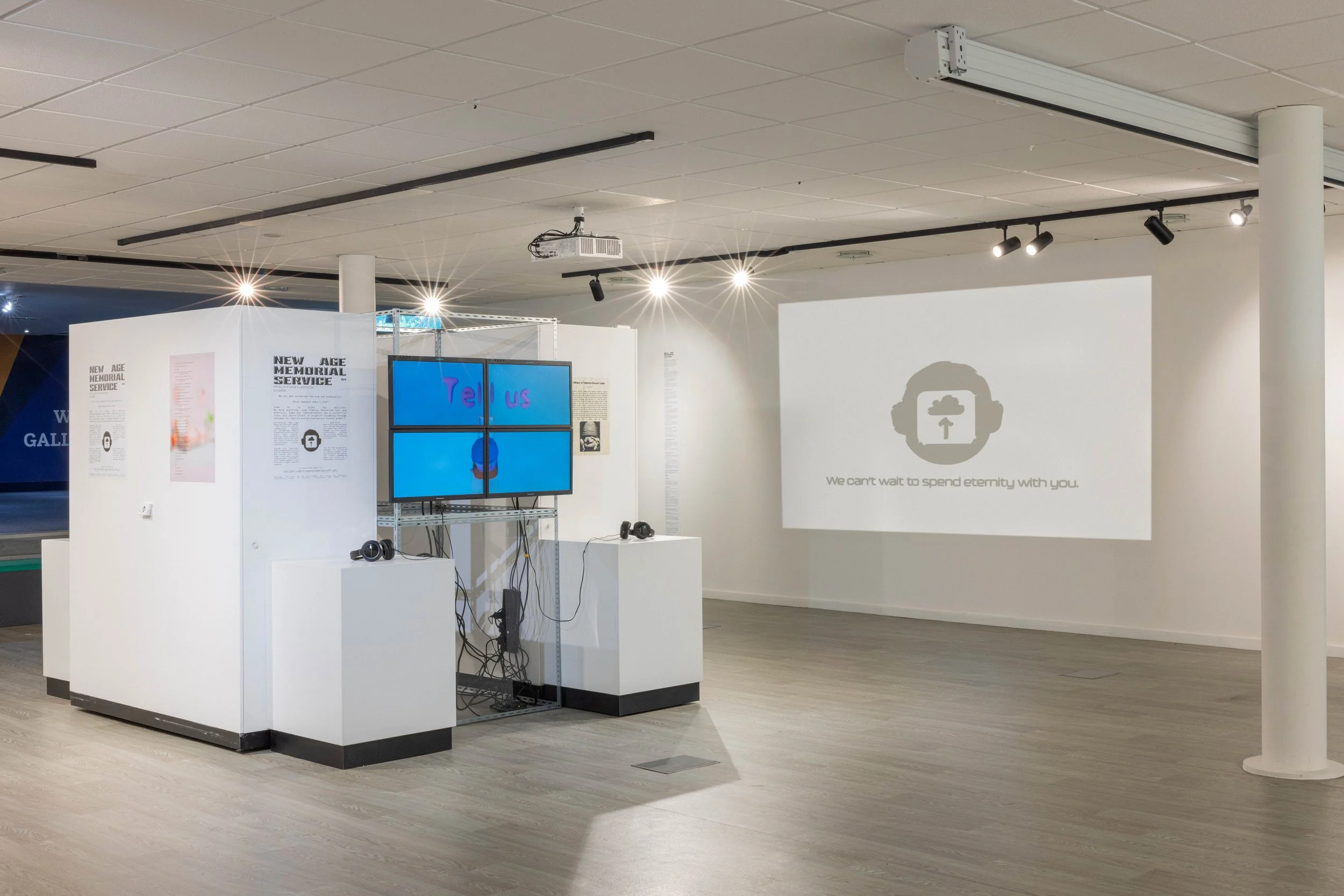 An art exhibit with white walls and floors, featuring a display with two screens showing the word "Tell us," and a projection on the wall with a stylized face wearing headphones and text that says, "We can't wait to spend eternity with you."
