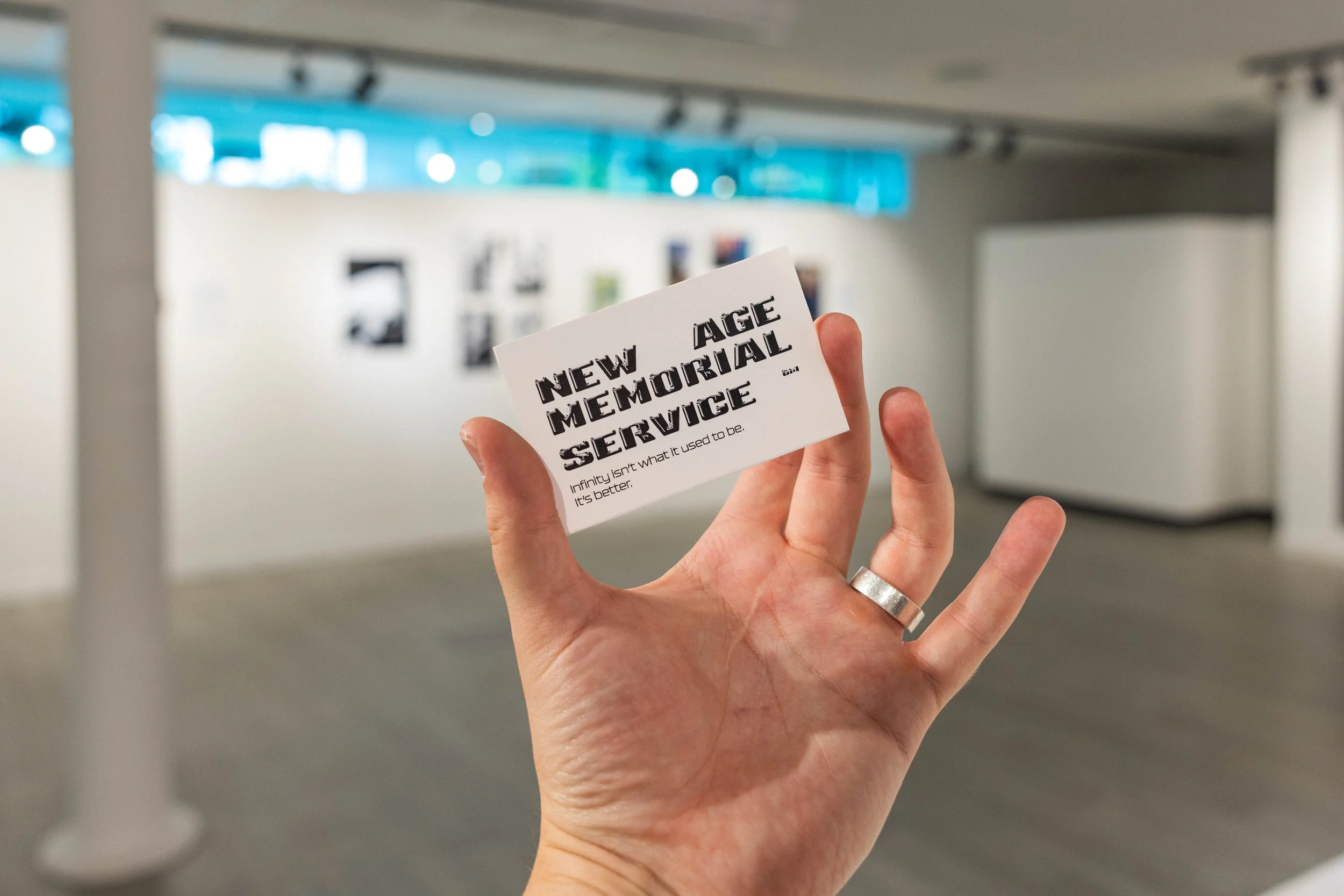 A hand holding a business card that reads 'New Age Memorial Service' with the subtitle 'Infinity isn't what it used to be. It's better.' The background shows an art gallery with white walls and framed pictures.