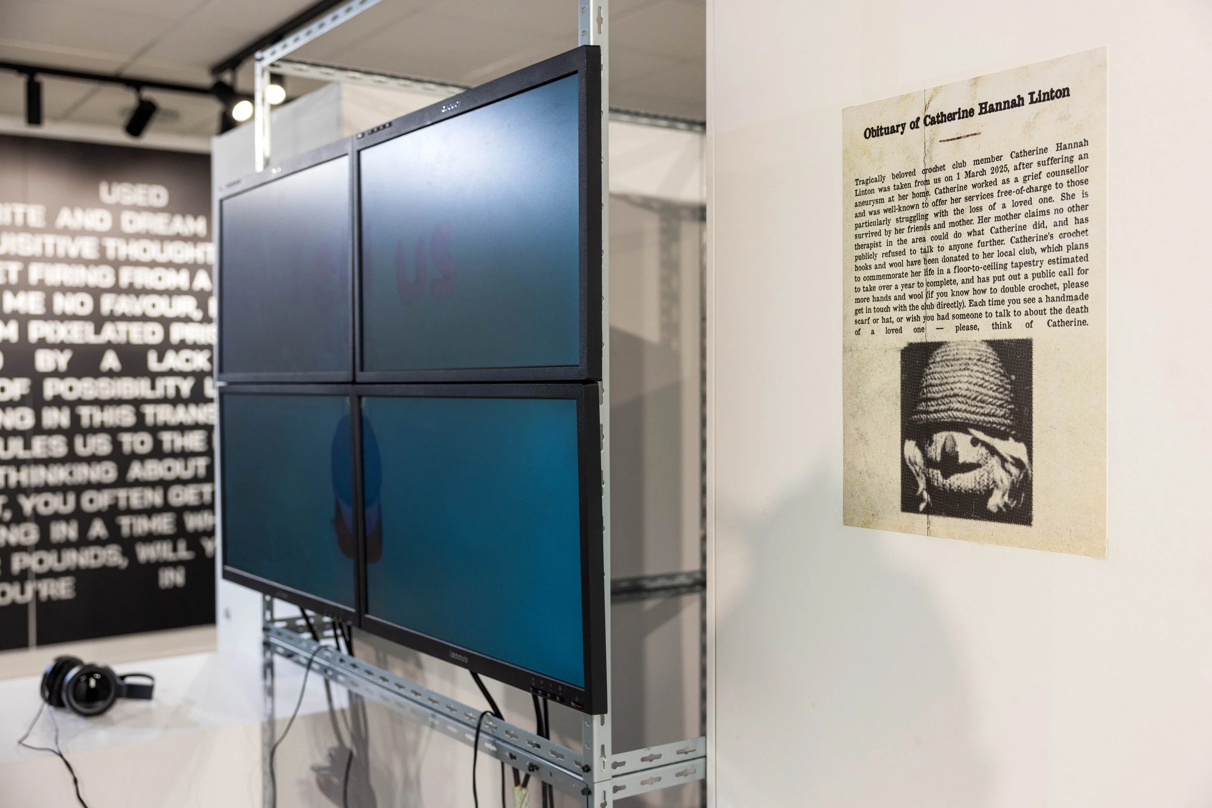 An art exhibit featuring a wall with a printed obituary for Catherine Hannah Linton and a photograph of a digital character. To the left, a large TV displays a Digital Film, and headphones rest on a table.