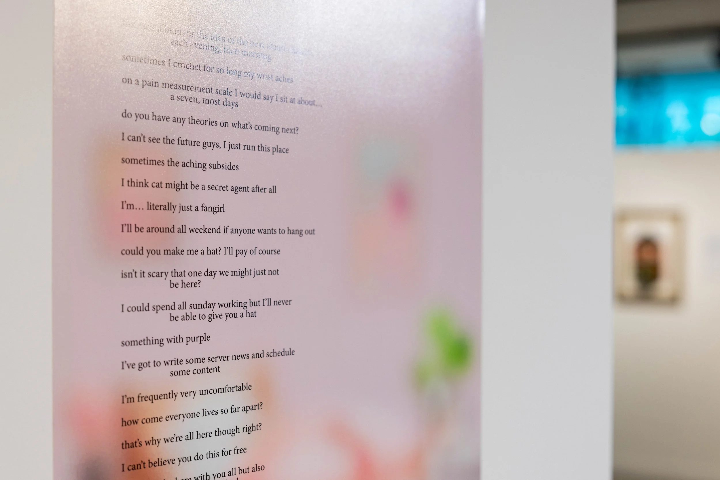 A close-up of a printed poem or song lyrics on a pink background, with the background slightly out of focus and hints of green, red, and purple. A blurred interior space is visible in the distance.