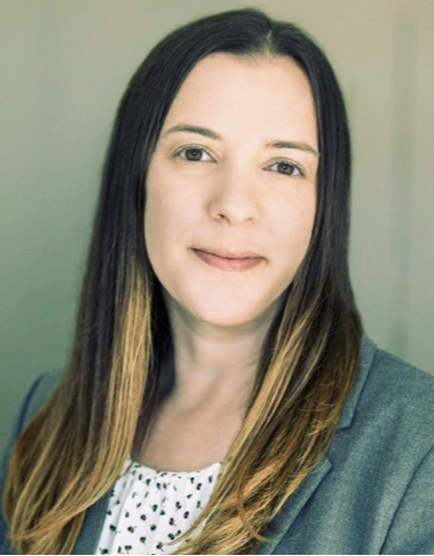 A woman with long, straight, dark brown hair and light skin, wearing a gray blazer over a polka-dotted blouse, smiling softly in front of a neutral background.