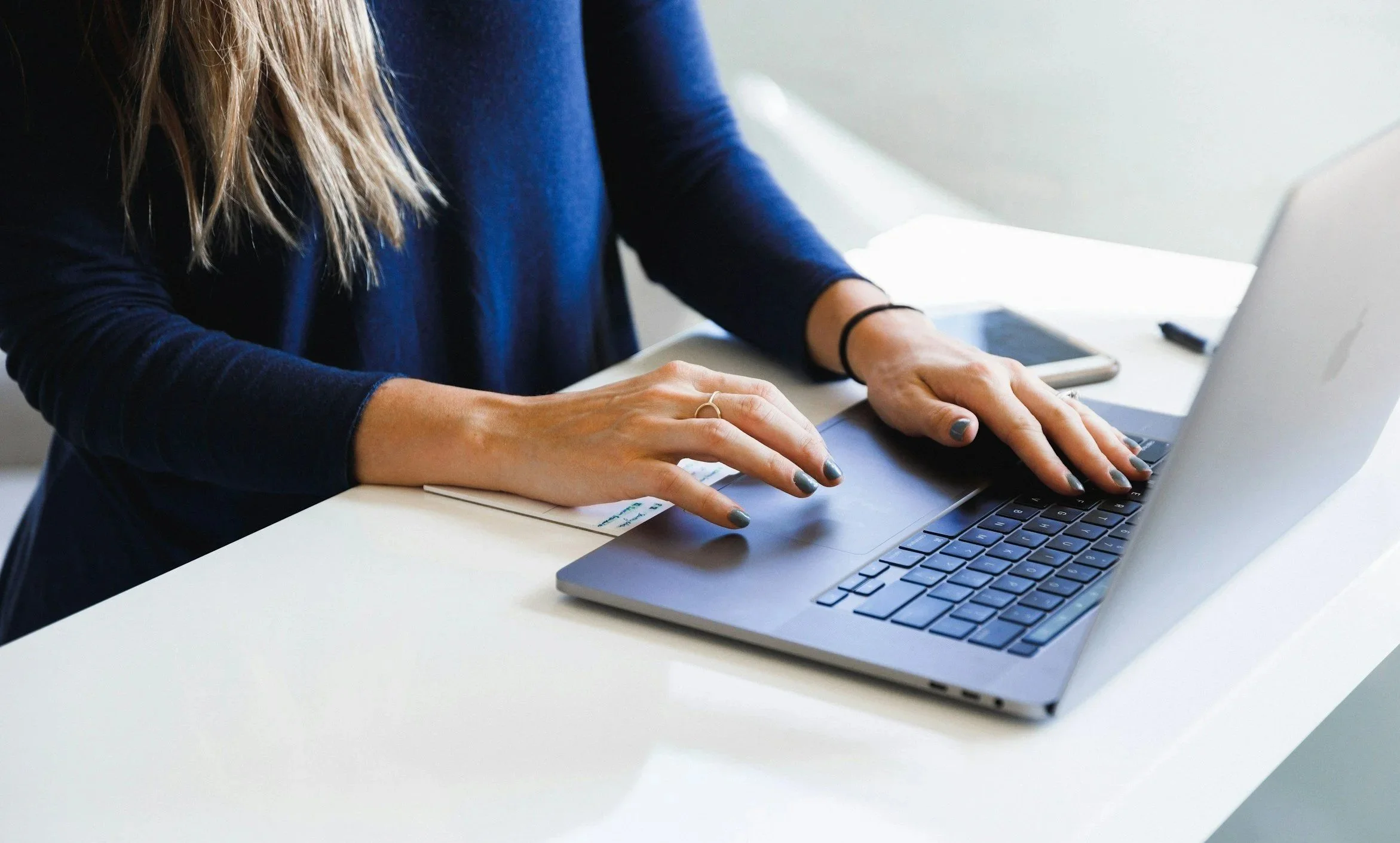 A woman with painted nails wearing a dark long-sleeve shirt using a laptop on a white table.