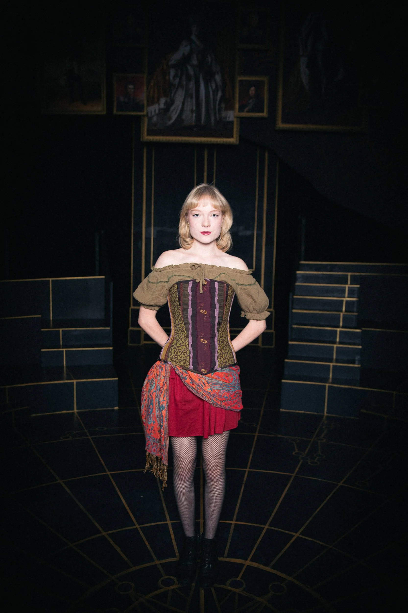 Actress and performer Olivia Scott, a young woman with blonde hair standing in a dark room with several portraits on the wall behind her, dressed in a colorful Victorian-style outfit, in Natasha, Pierre, and the Great Comet of 1812 production.