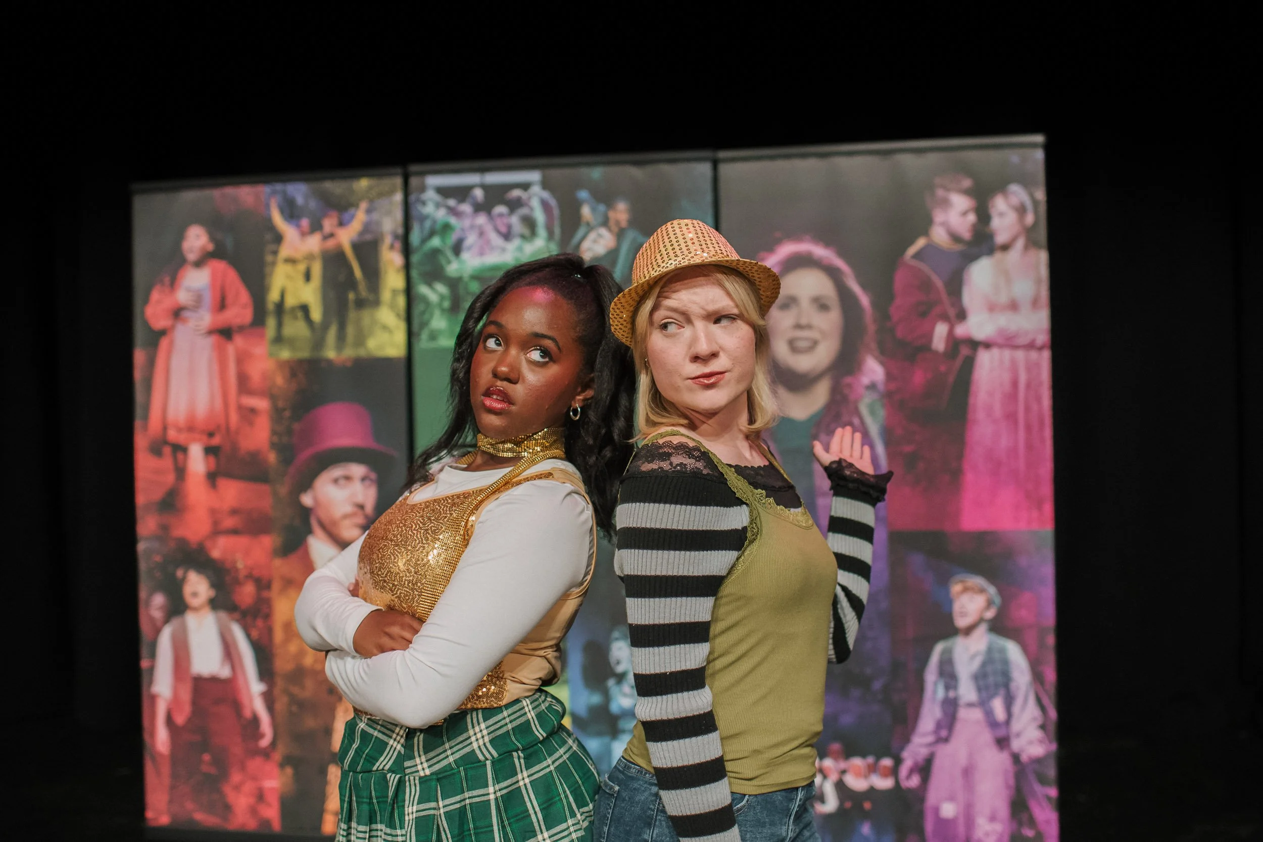 Two women stand back-to-back on stage, looking skeptical. They are in front of a colorful backdrop with various theatrical images.