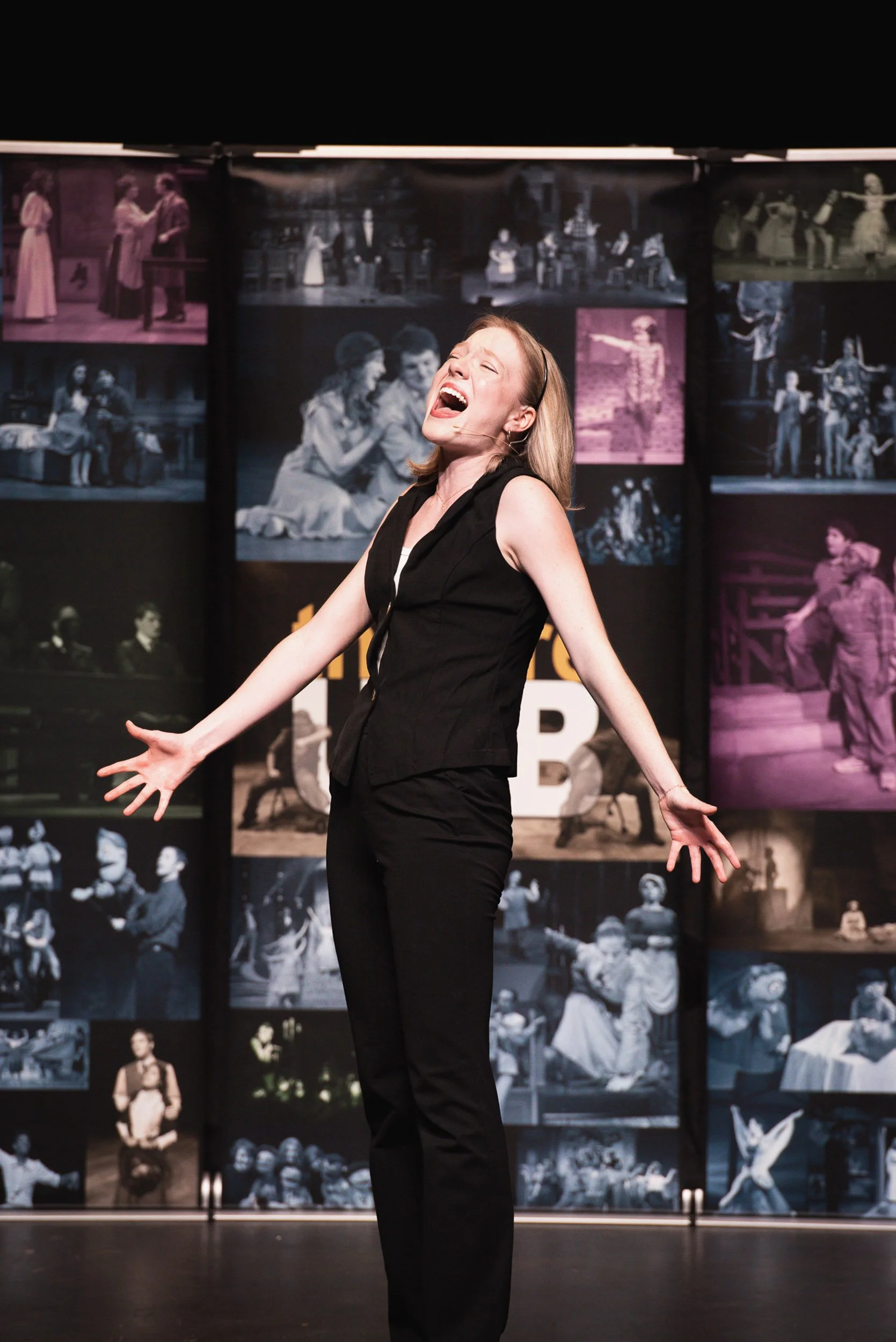 A woman in a black sleeveless top and black pants is on stage with her arms outstretched and head tilted back, singing or performing with a joyful expression. Behind her is a backdrop filled with black-and-white photos of theatrical performances and 