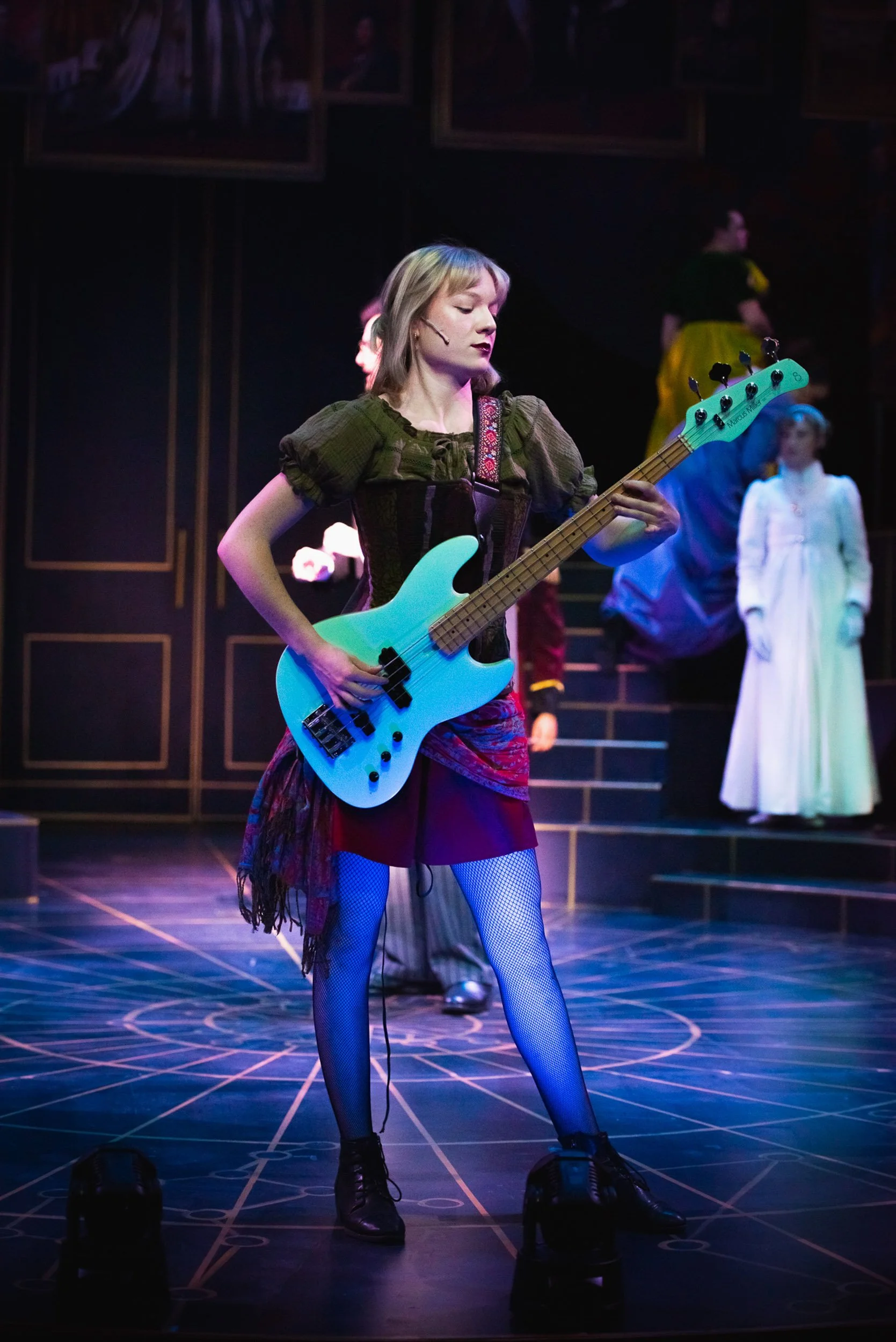 Actress and Musical Theatre Performer Olivia Scott is playing an electric bass guitar on stage during a theatrical performance, with other actors in period costumes in the background in the production Natasha, Pierre, and the Great Comet of 1812.
