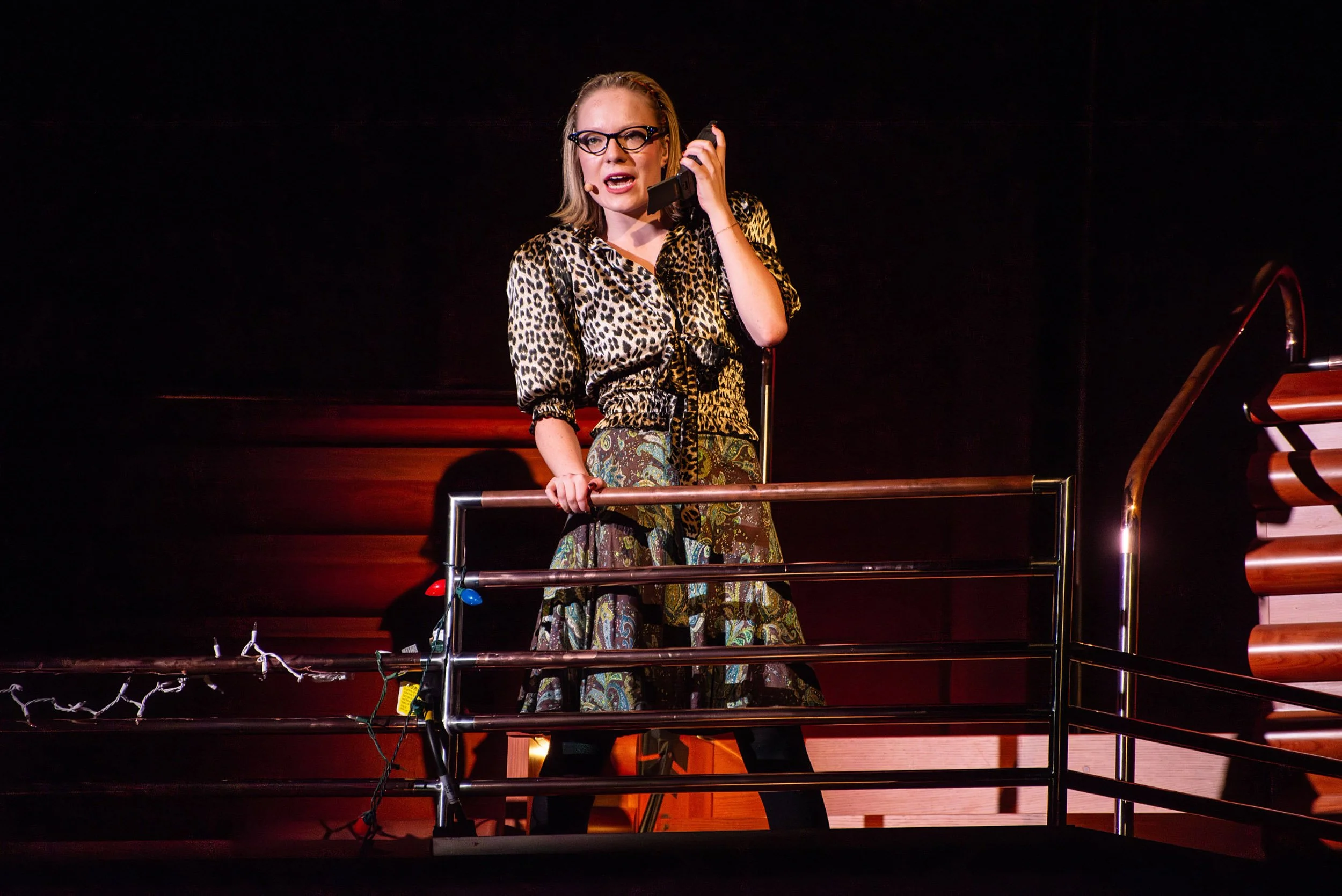 Young actress Olivia Scott with glasses and leopard print blouse speaking into a headset microphone on a stage, standing behind a railing with Christmas lights in RENT production.
