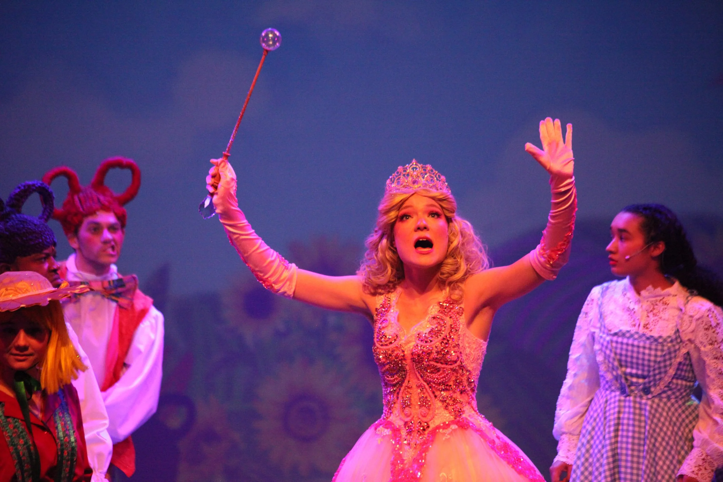 A scene from a theatrical performance featuring dressed as a princess in a pink ball gown, wearing a tiara, holding a scepter, with an expression of surprise or shouting. She is surrounded by actors in colorful costumes, including characters with ani