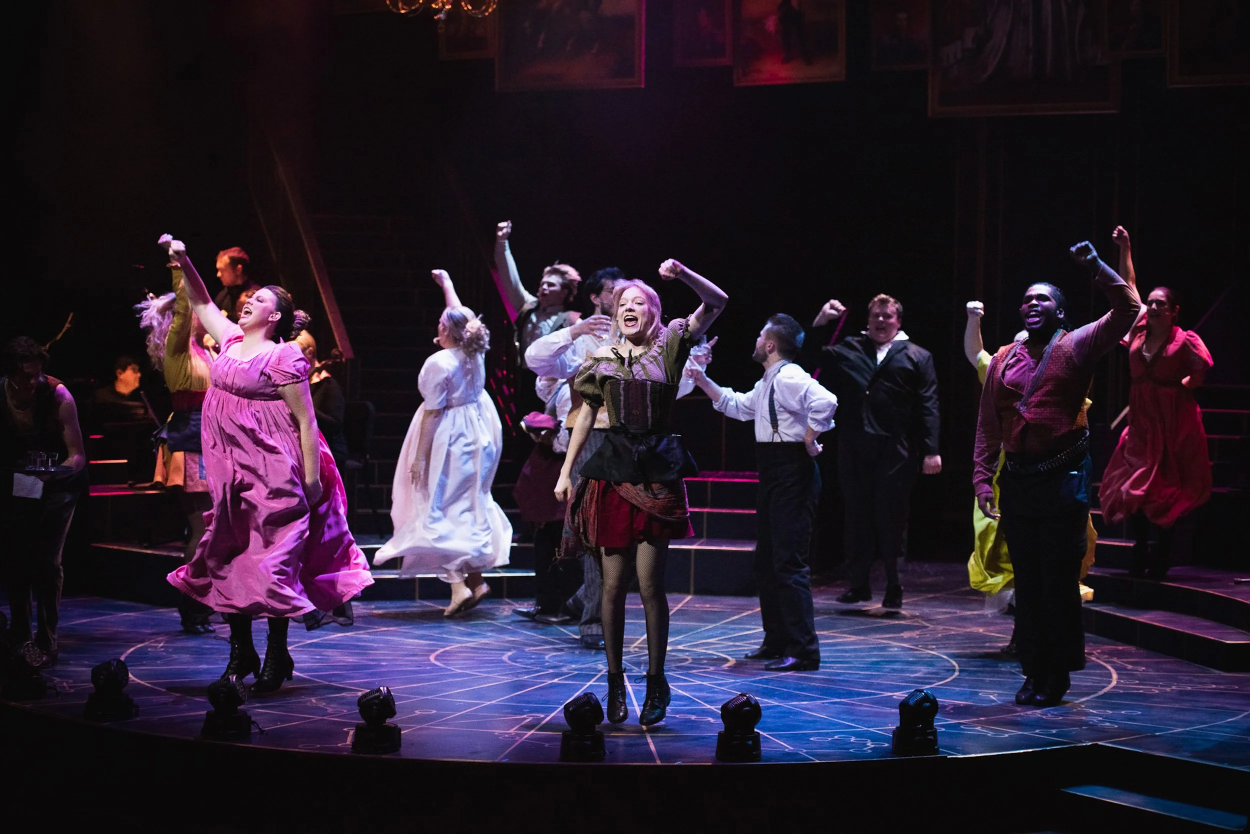 Actors performing a dance on a stage with colorful costumes, with Olivia Scott, actress and performer, with raised fists, in a theatrical production.