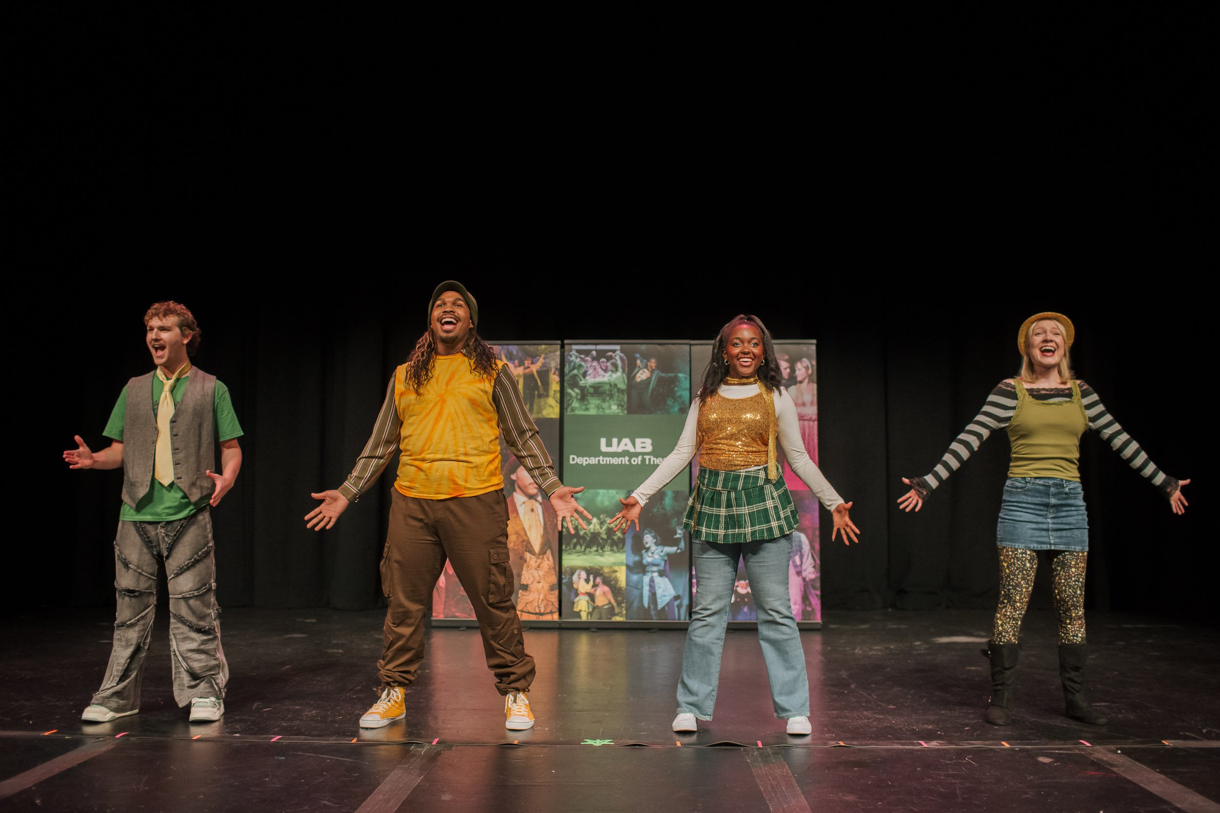 Four performers on stage during a theatrical production, smiling and singing, with a UAB Department of The Arts banner in the background.