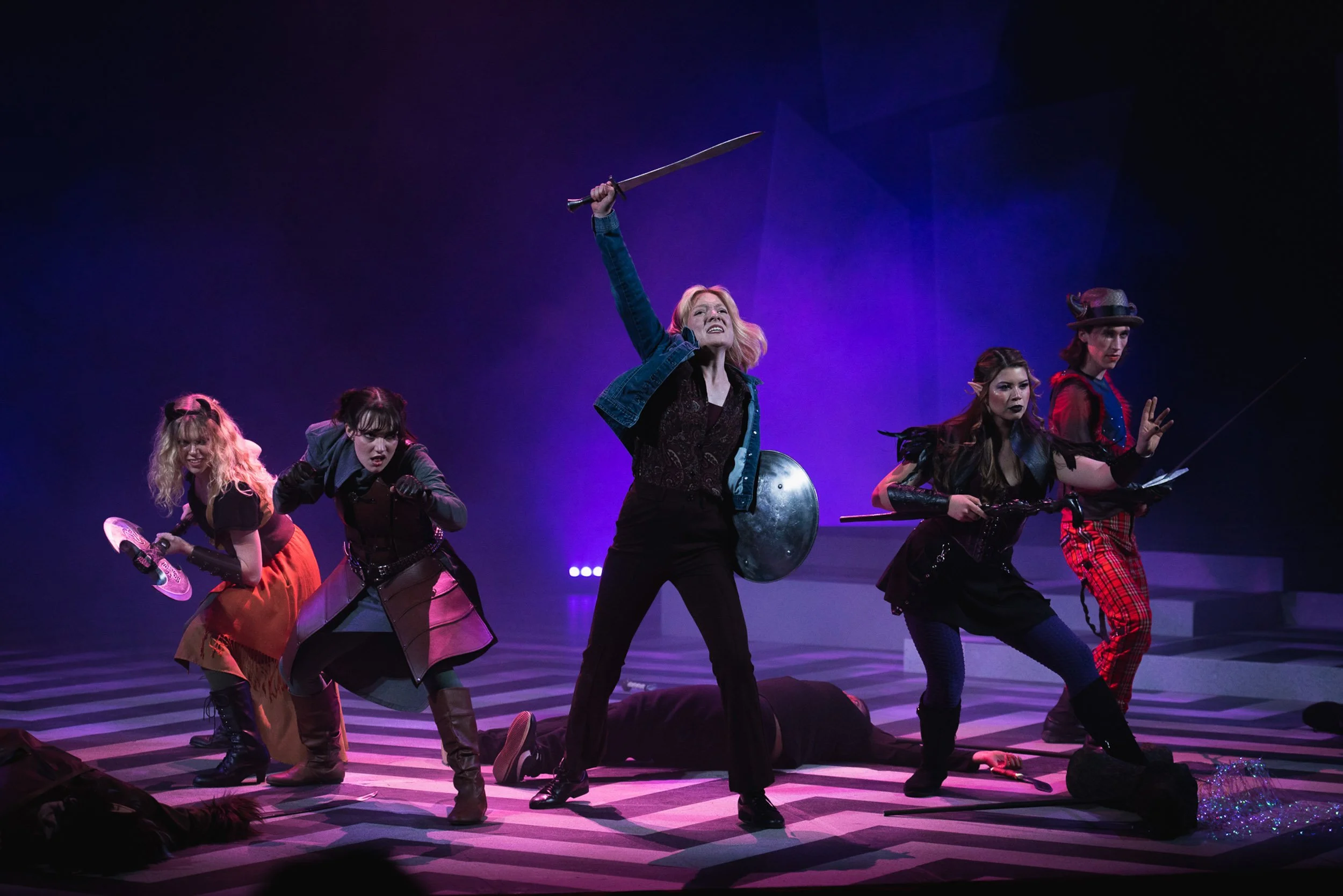 Dramatic scene from a theatrical performance featuring actress Olivia Scott and four other women in costume, with one woman standing in the center raising a sword and holding a shield, others in various poses, on a stage with purple lighting.