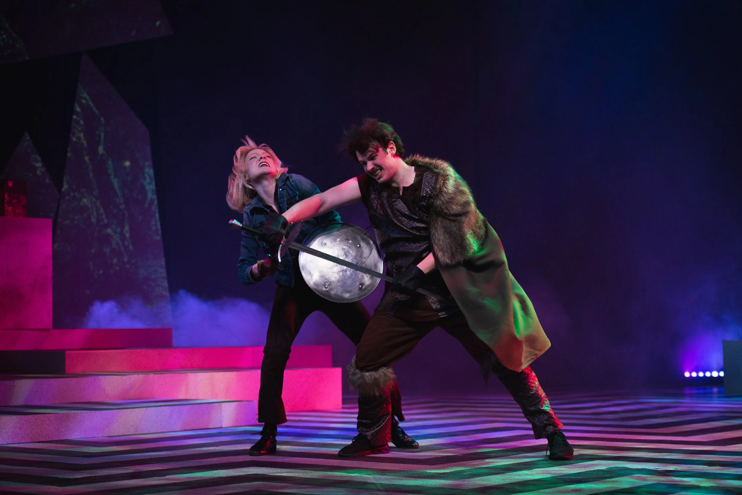 Two actors performing a dramatic fight scene on stage, Actress and entertainer Olivia Scott with a shield and a spear, the other in a fur-trimmed cape, with colorful lighting and a dark backdrop.