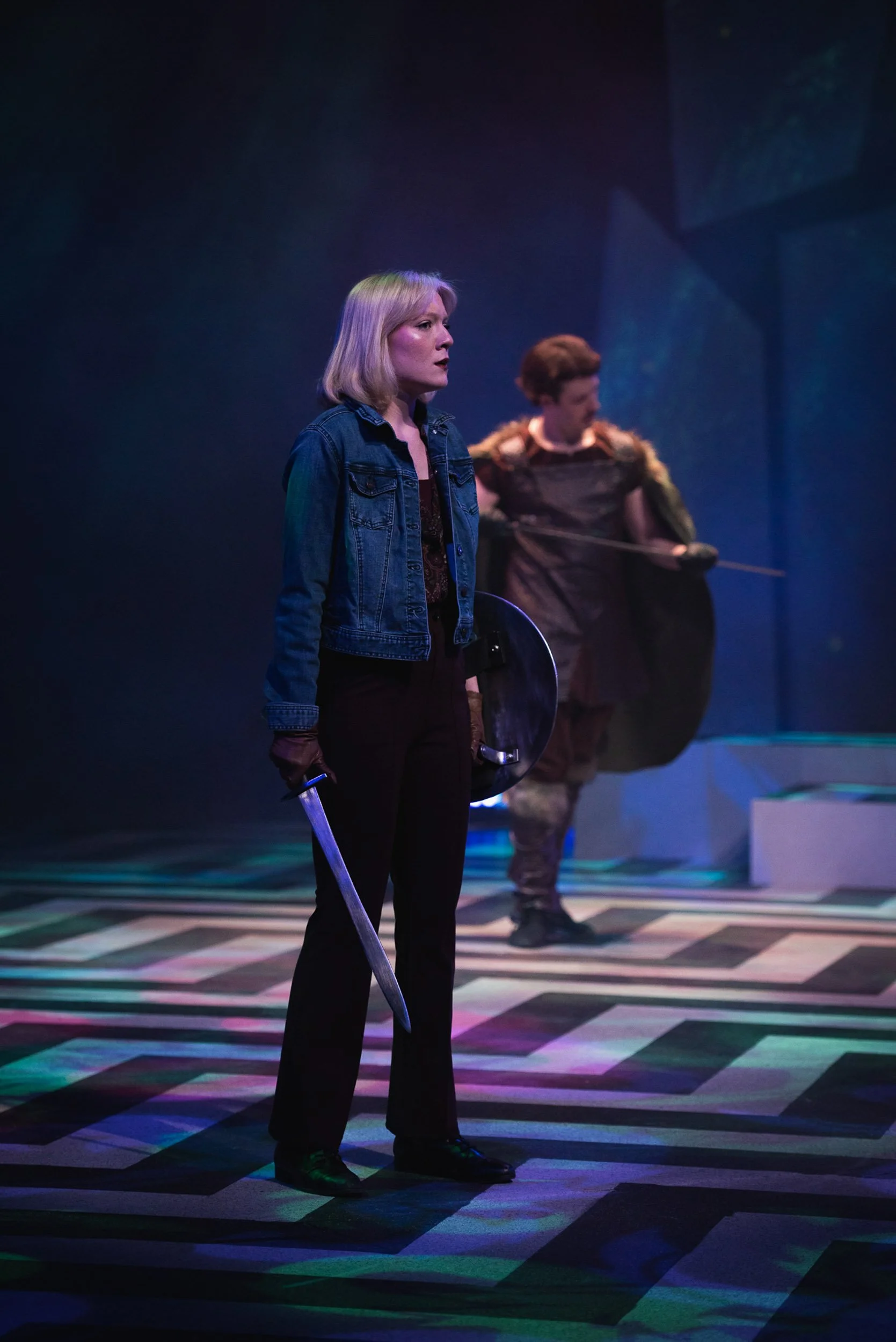 A scene from a theatrical play with two actors on stage. The woman in the foreground has blonde hair, holds a sword and a shield, and wears a denim jacket and black pants. The man in the background wears medieval armor, holding a spear, with a dark c