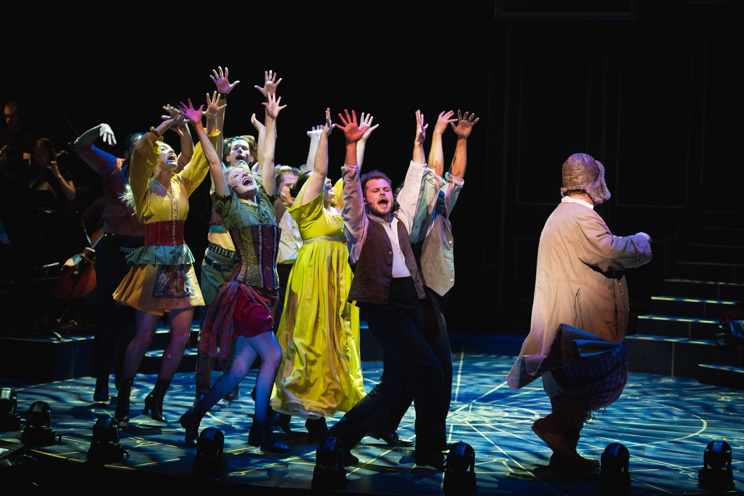 Theater cast performing on stage with their hands raised, dressed in colorful period costumes, and an orchestra in the background.