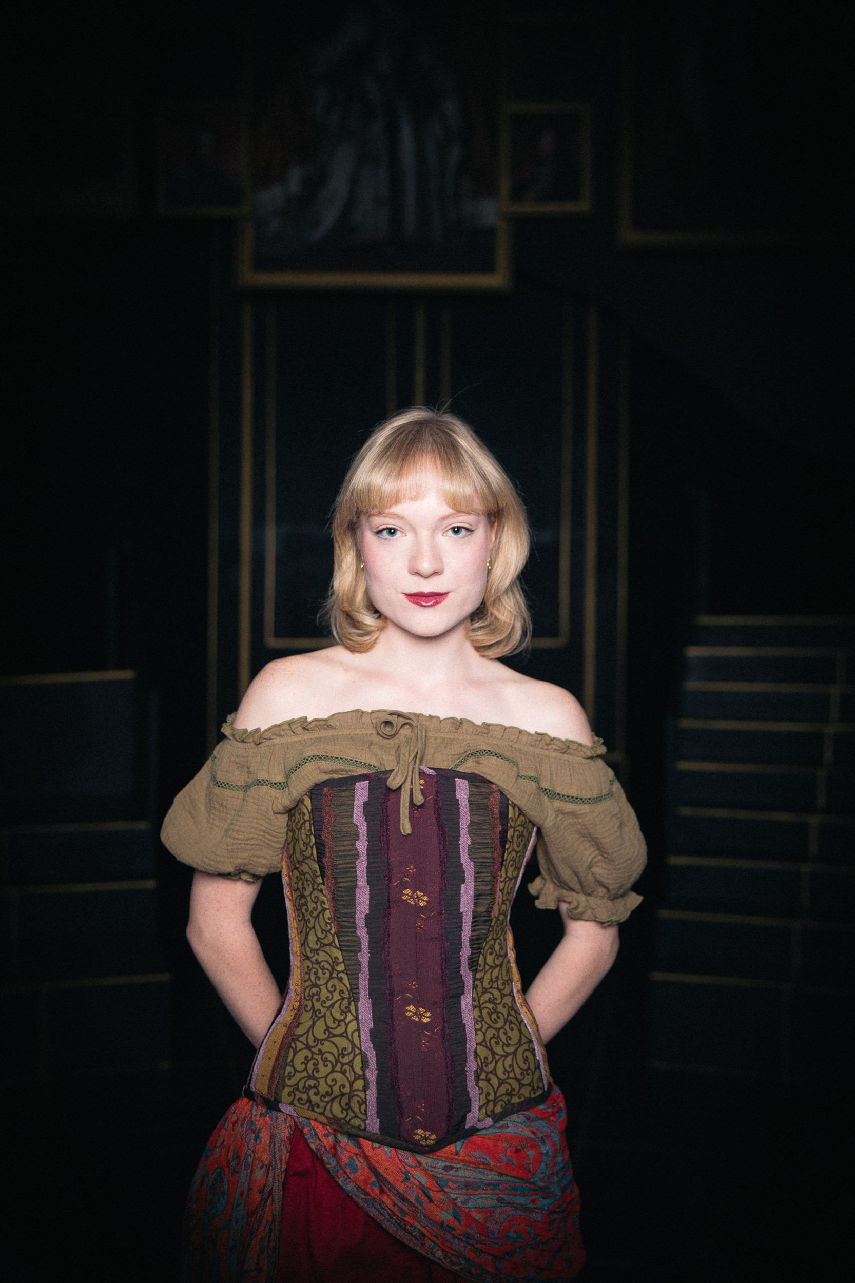 Actress and performer Olivia Scott with blonde hair in a colorful corset and off-the-shoulder top standing in a dark room with paintings in the background.
