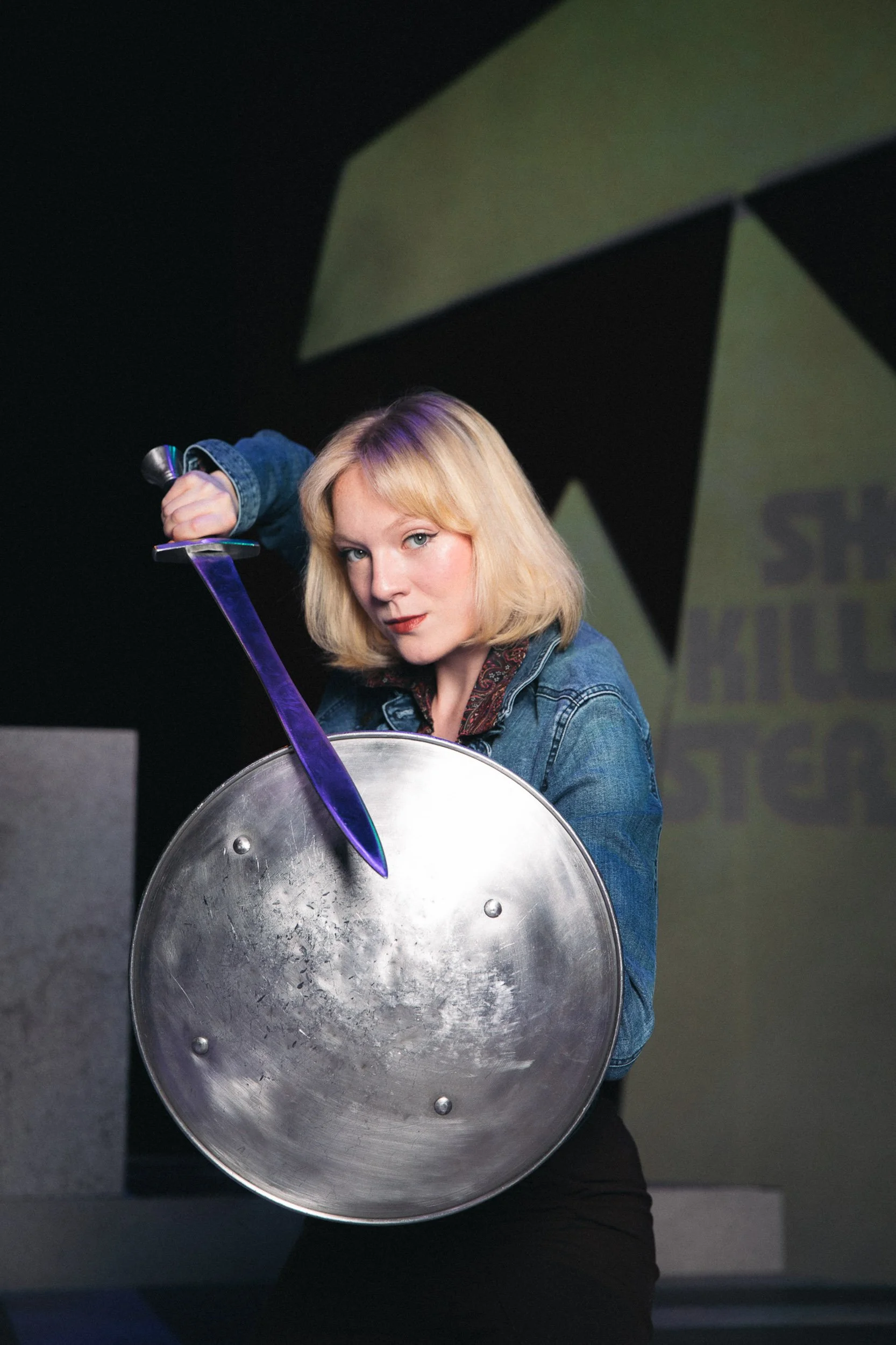 Actress and entertainer Olivia Scott with blonde hair in a denim jacket holding a sword and a shield at a stage performance or event.