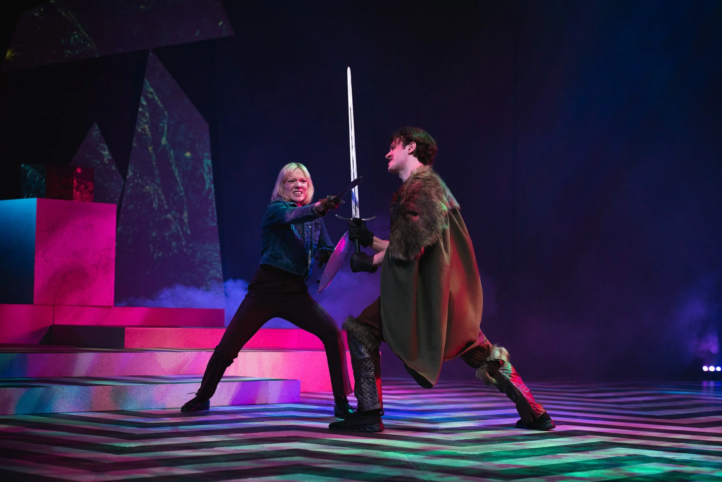 Actress and entertainer Olivia Scott and another actor perform a theatrical battle scene on stage, with one woman wielding a sword and shield and a man dressed in medieval costume, kneeling with a sword