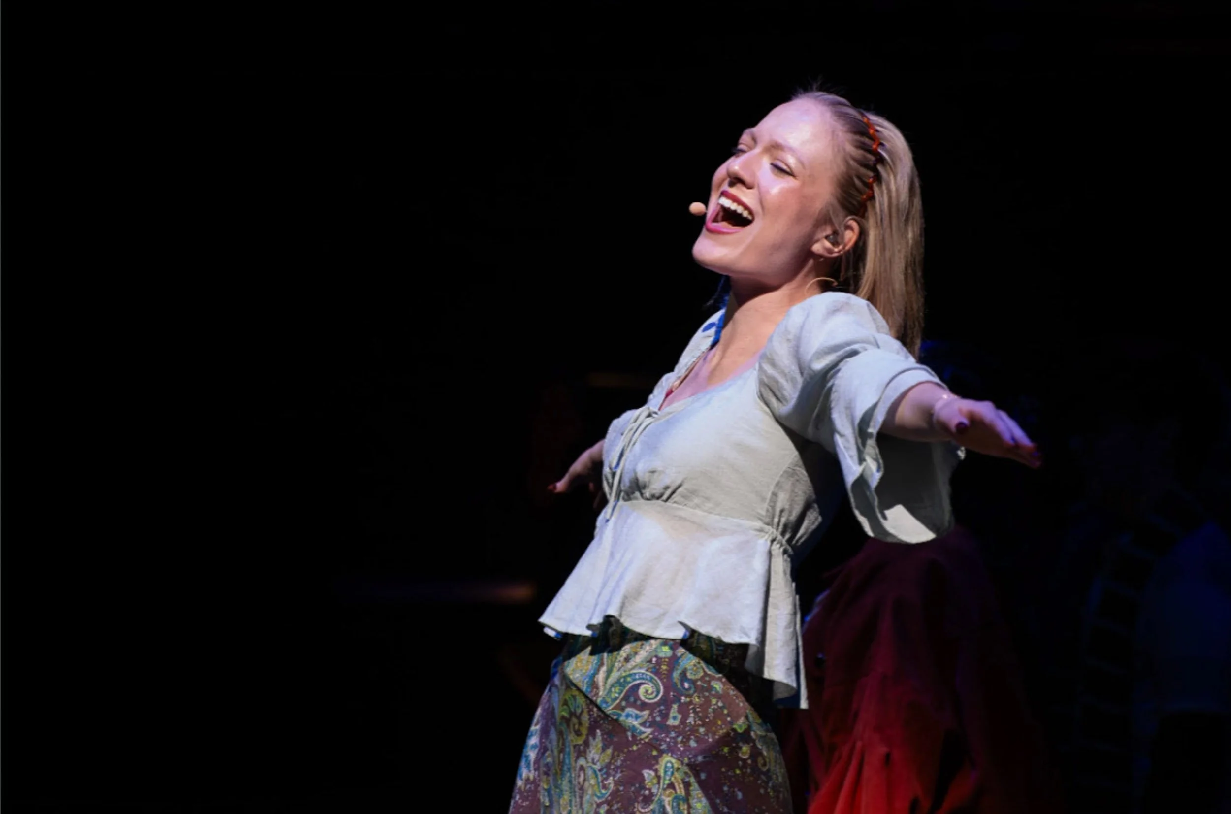Actress and singer Olivia Scott singing on stage with arms outstretched, wearing a light-colored blouse and patterned skirt, with a smile and closed eyes, illuminated by stage lighting, in the RENT play.