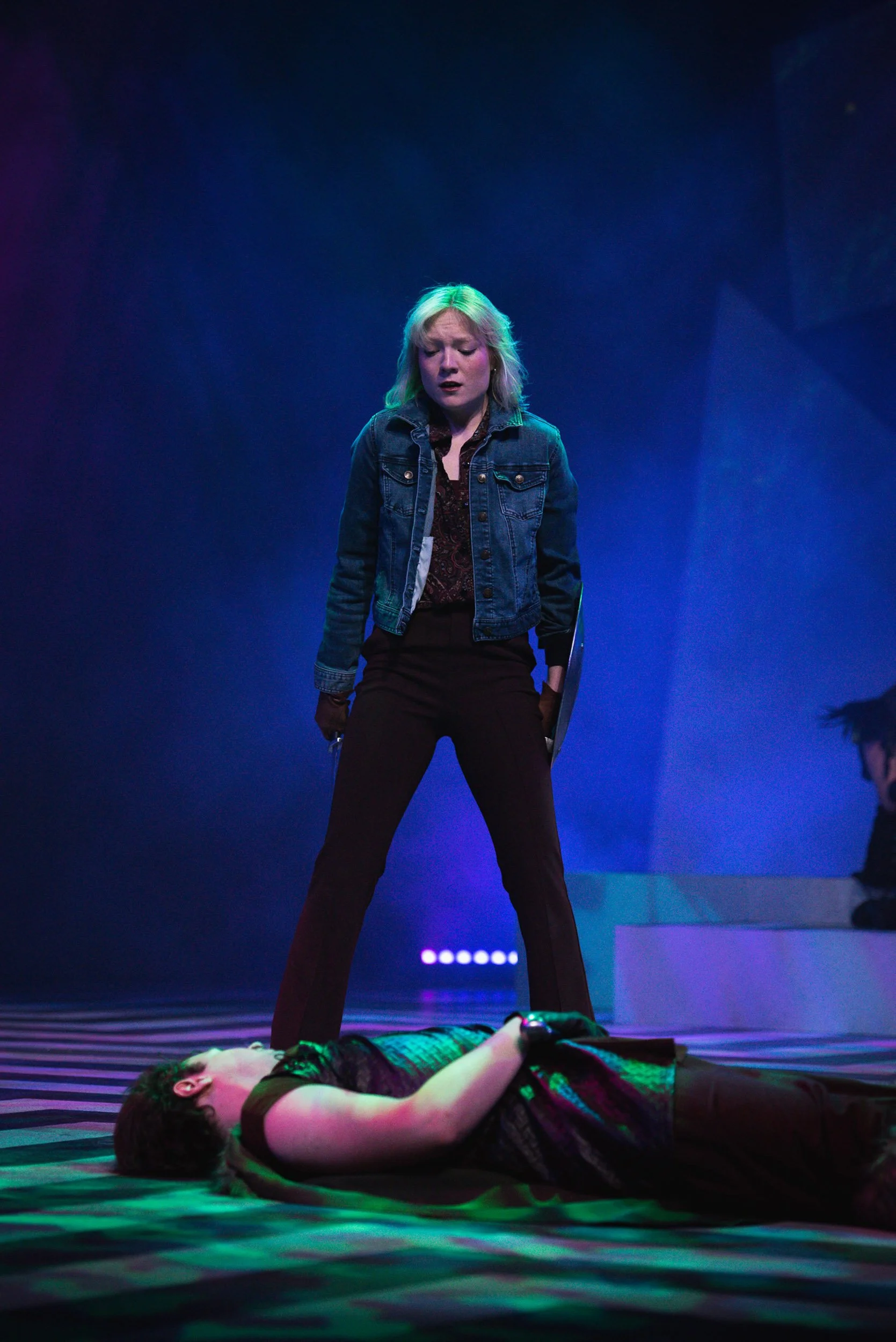 Actress and entertainer Olivia Scott with blue-green hair standing over a person lying on the floor during a theatrical performance, with dim blue and purple stage lighting.