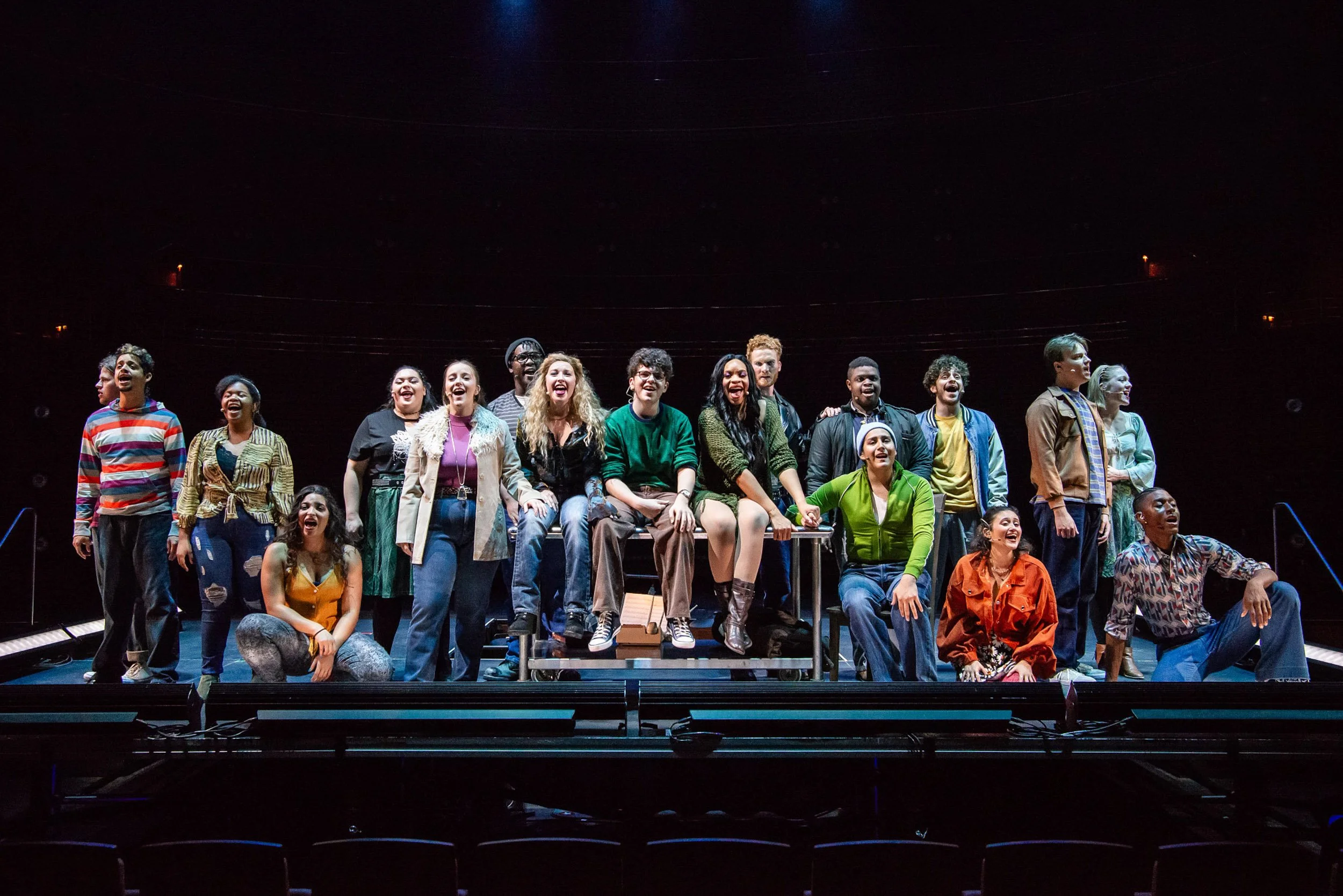 A diverse group of actors on stage during a theater performance, smiling and holding hands.