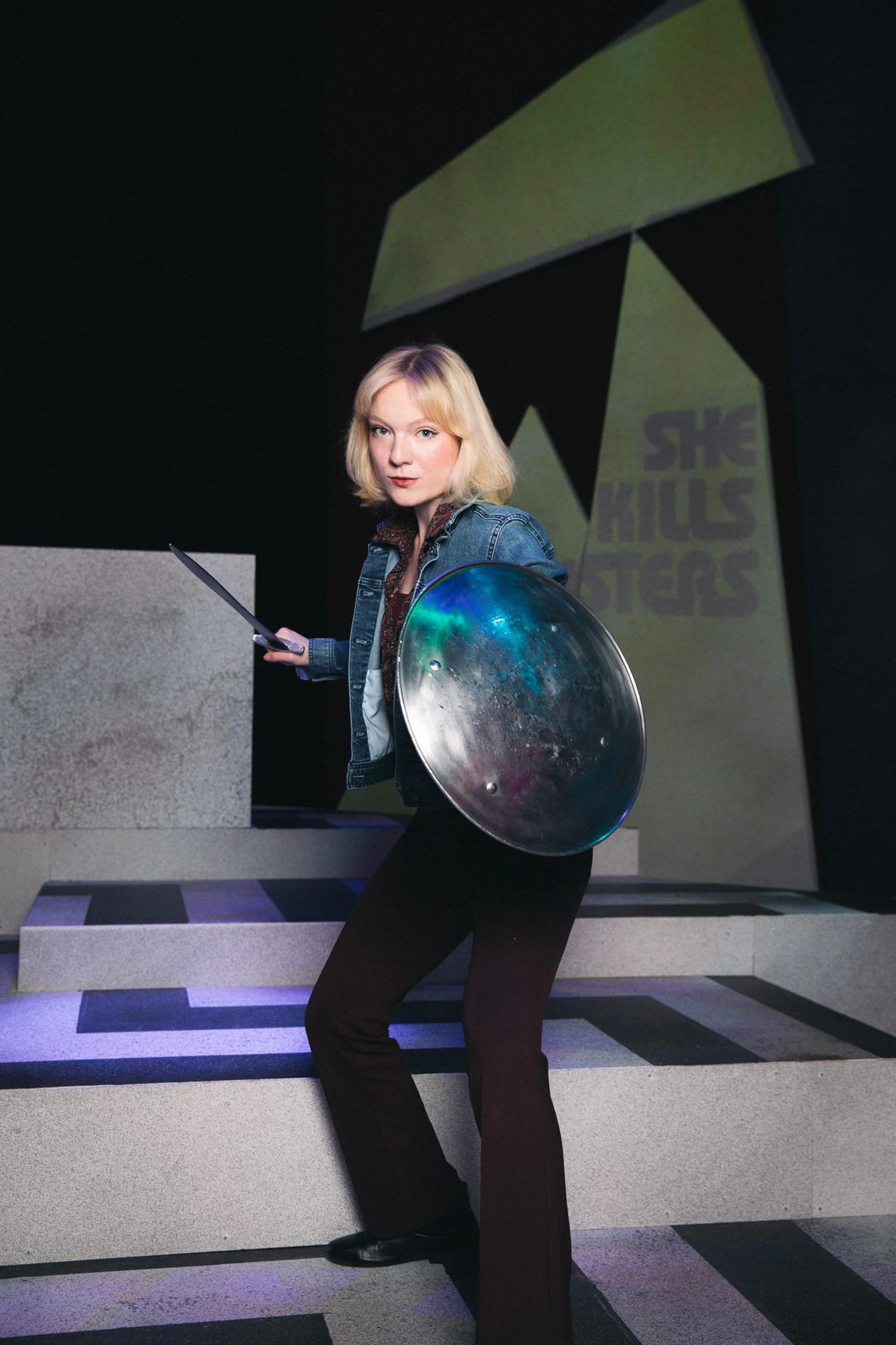 Actress and entertainer Olivia Scott with blonde hair in a denim jacket holding a sword and a shield stands on a set of stairs in a dimly lit environment, with a geometric background and a sign that reads 'SHE KILLS MONSTERS'.