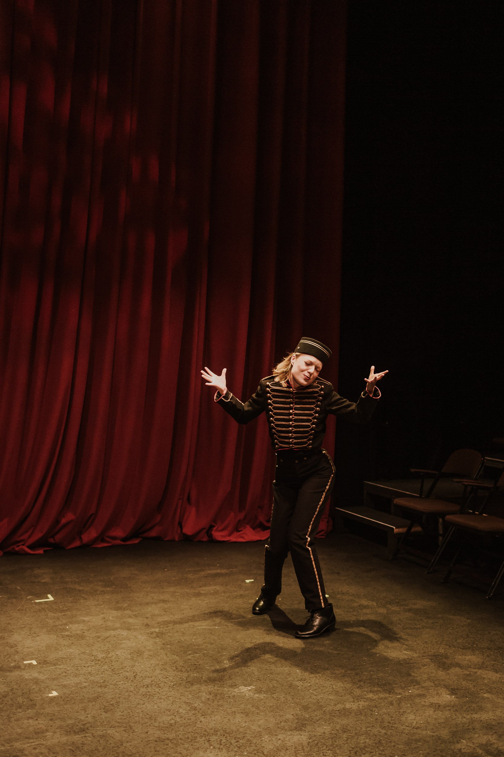 Olivia Scott, actress and performer  dressed in a black military-style jacket with gold trim and matching pants, performing a dance or theatrical pose on stage in front of red curtains.