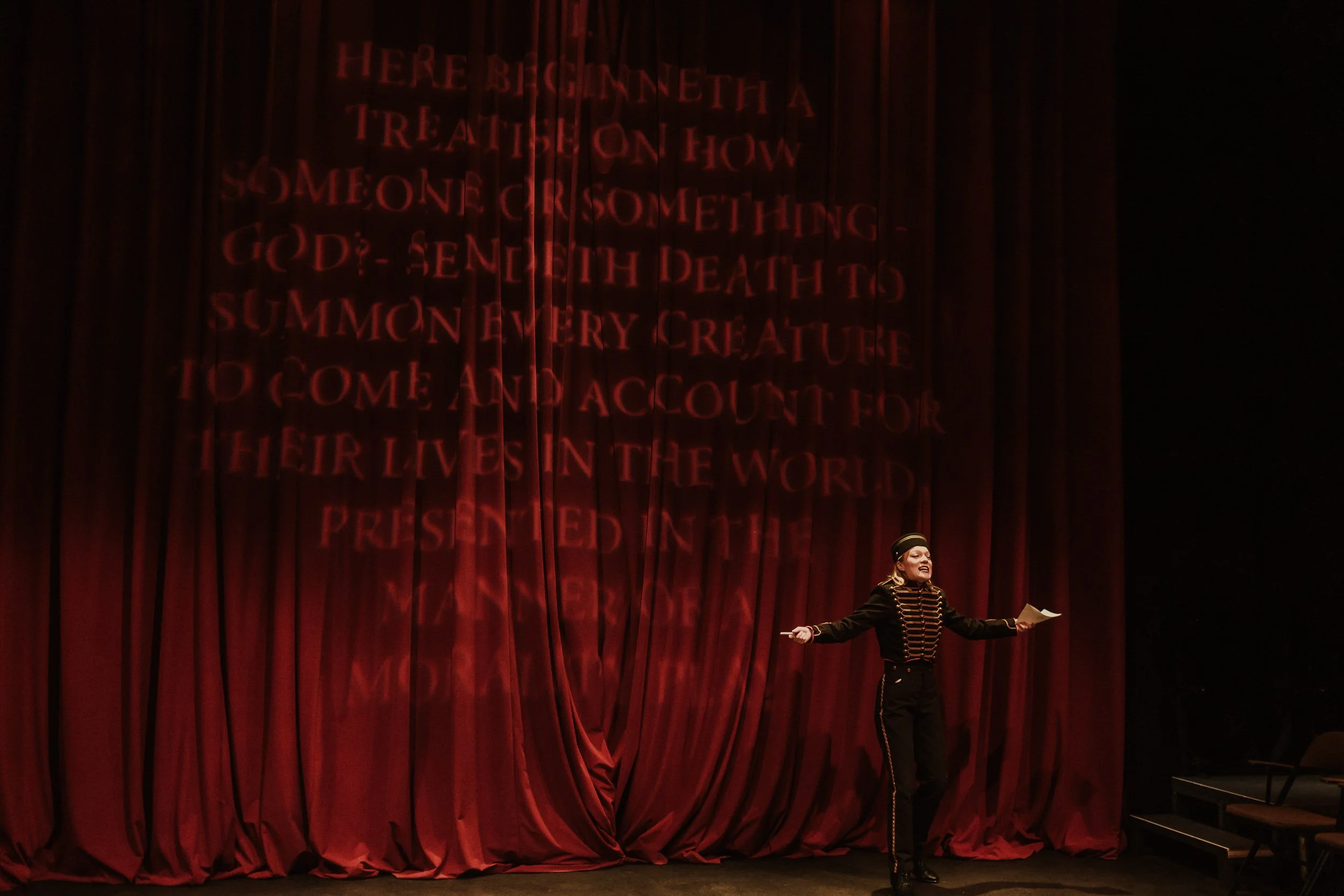 Olivia Scott, actress and performer on stage in a theatrical production, standing in front of a large red curtain with stage script projected on it, wearing a black military-style costume and holding papers in Everybody play.