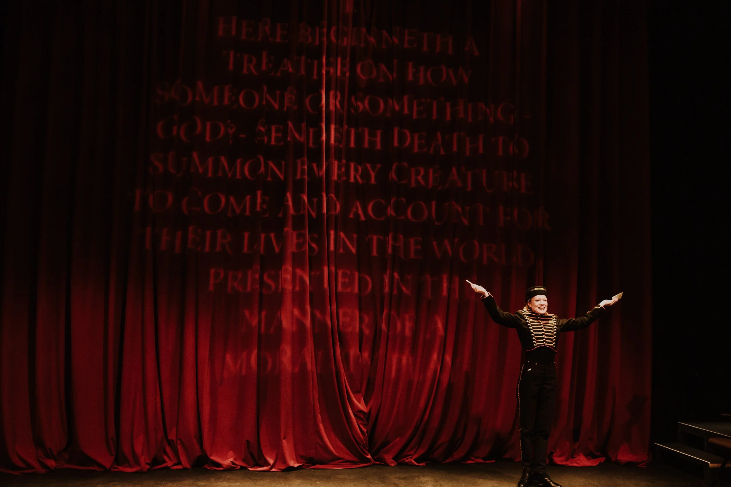 Olivia Scott, actress and performer in theatrical costume on stage with red curtain backdrop, arms outstretched, in her performance in the Everybody play.