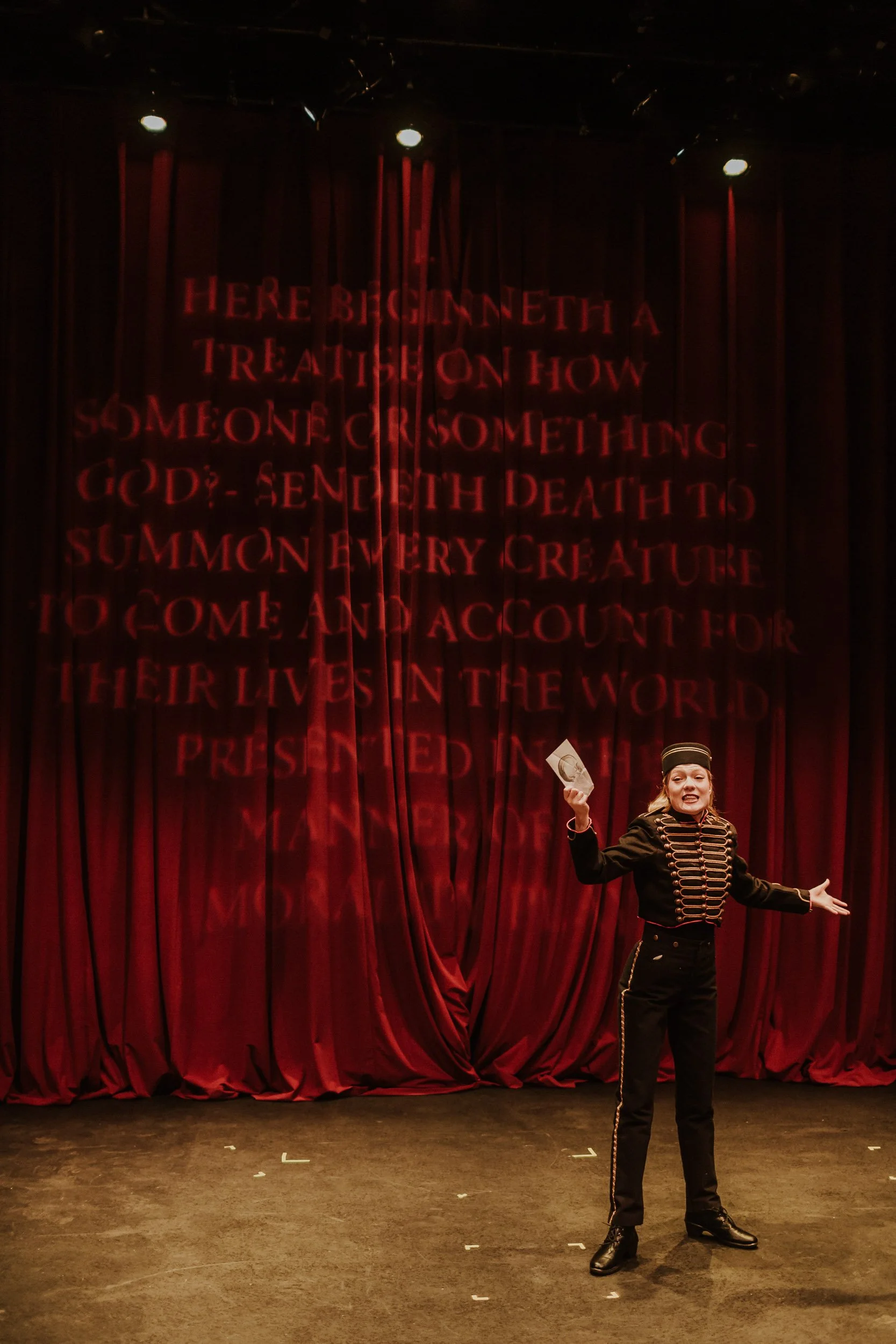 Olivia Scott, actress and performer dressed in a black uniform with gold buttons and a black hat, standing on a stage with red curtains, holding a piece of paper, and gesturing with arms open in her performance in Everybody.