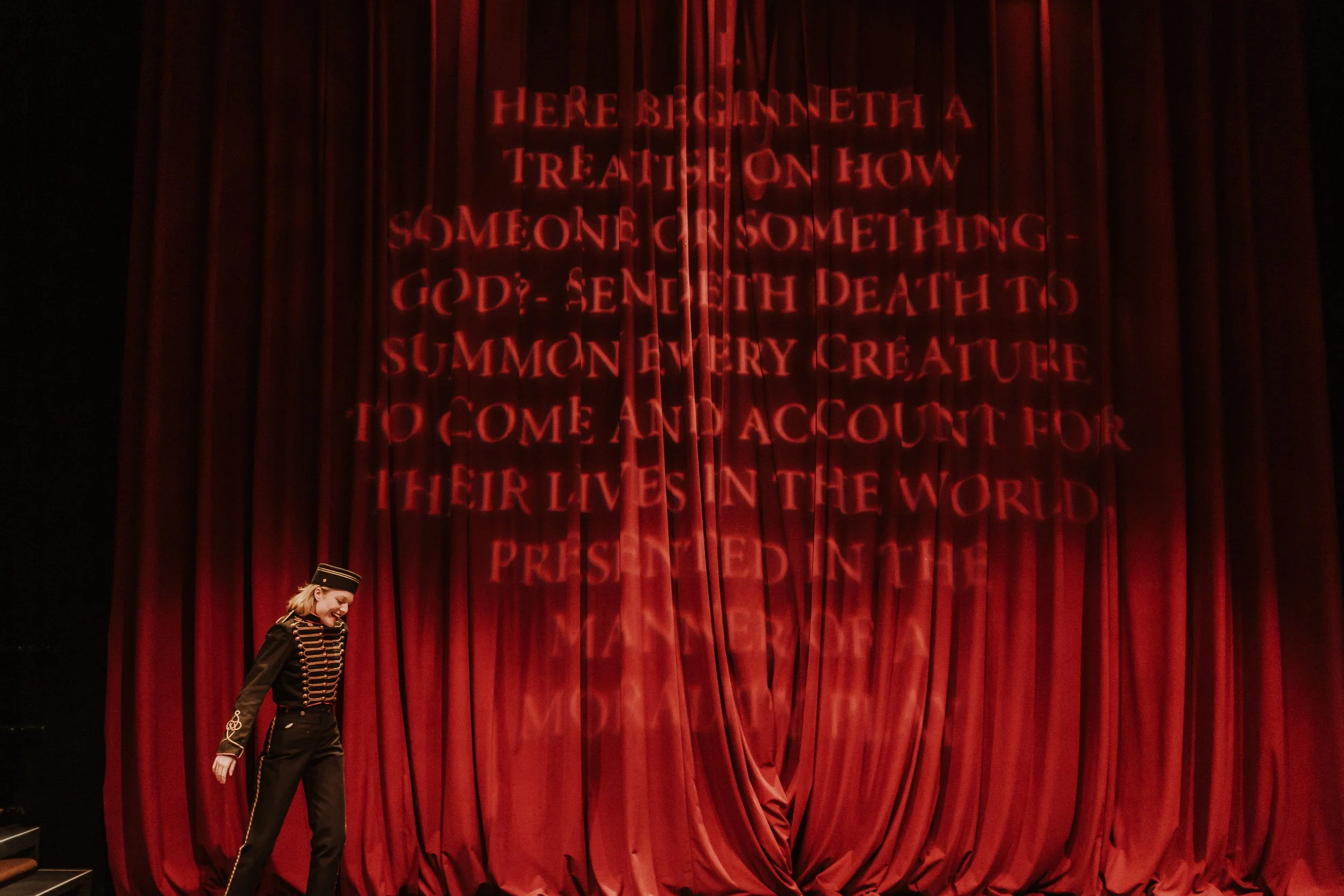 Olivia Scott, actress and performer dressed as a military band member standing on stage with a red curtain behind and a message projected onto it in her performance in Everybody. 