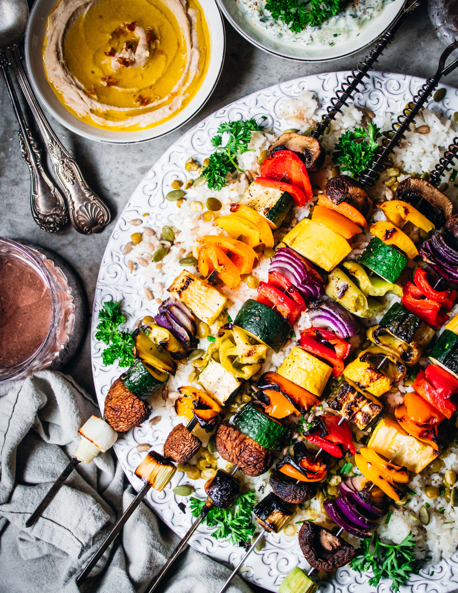 Colorful vegetable and tofu skewers on a white plate with rice, garnished with parsley and seeds, with dips or sauces in bowls nearby.