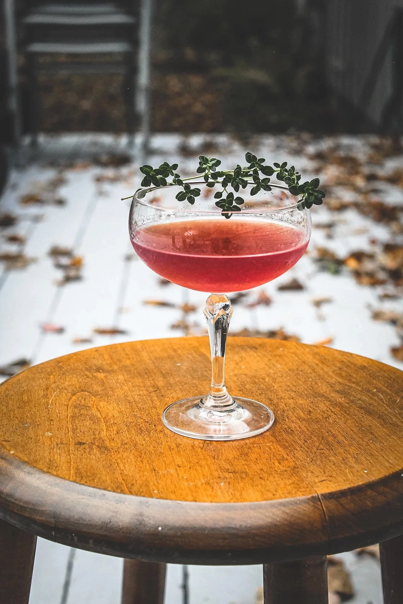 A pink cocktail in a coupe glass garnished with a sprig of fresh thyme, placed on a round wooden table outdoors with fallen autumn leaves.